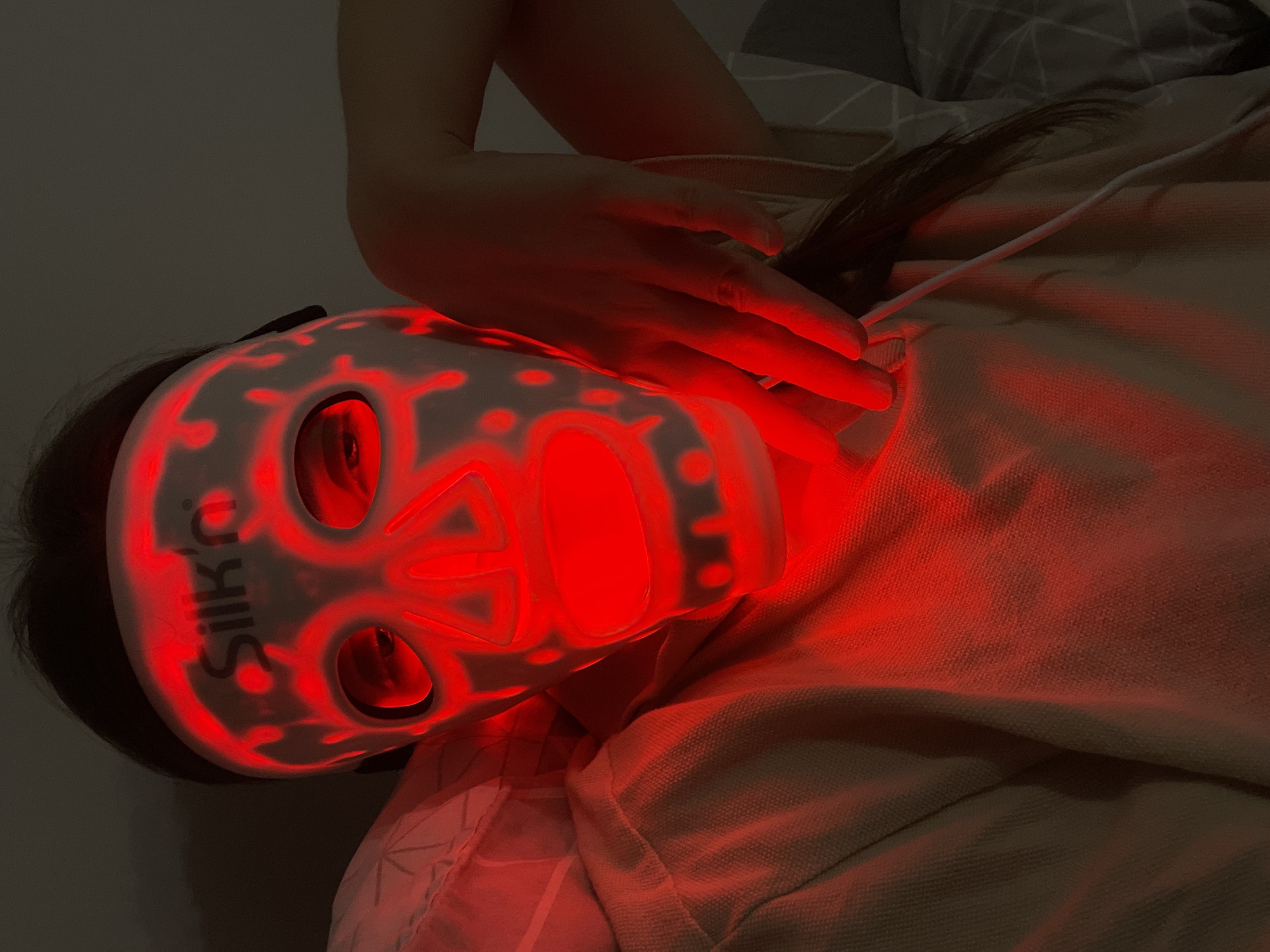 A person is lying on their back with a red LED face mask on, identified by the "Silk'n" logo. Their hand is placed on their neck. A white wire connects to the mask. The person has dark hair, and they are on a bed with a patterned pillow. Location: Hévíz, Hungary is not evident in the image.
