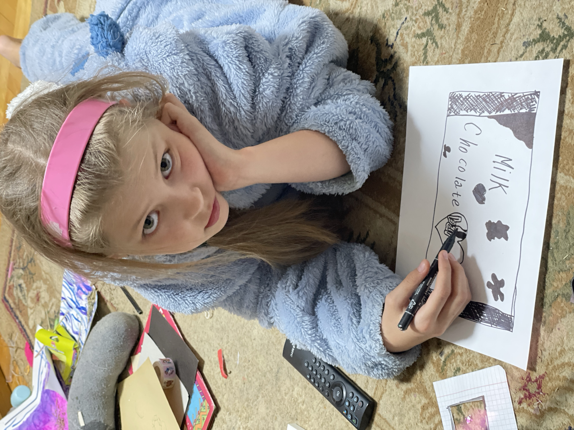 A girl with blond hair and a pink headband, wearing a light blue fuzzy robe, is lying on a patterned carpet. She is drawing a picture of a milk chocolate bar on white paper with a black marker. The drawing includes the words "MILK" and "Chocolate." To the left of the girl are various craft supplies, including a gray sock, paper, and tape. A black remote control is also visible. The scene is inside a home. There is no information within the image indicating that the location is Izhevsk, Russia.