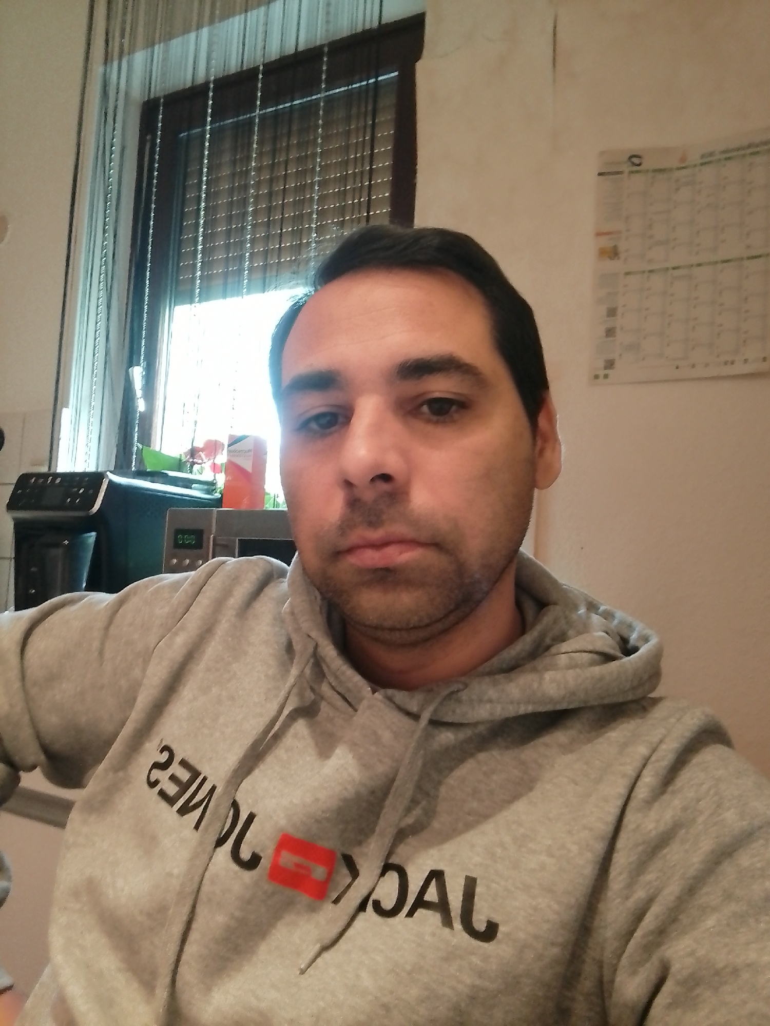 A man, with short, dark hair and stubble, is taking a selfie in a room. He is wearing a grey hooded sweatshirt with black lettering that appears to say "JACK & JONES" with a red rectangular logo attached to the front. In the background, on the left, is a window with vertical blinds and a sheer curtain. Next to the window is a black appliance, possibly a coffee maker, and a silver microwave. On the right side of the background is a wall calendar. The room appears to be domestic, possibly a kitchen or living area. There is no specific indication of the location within Delmenhorst, Germany.