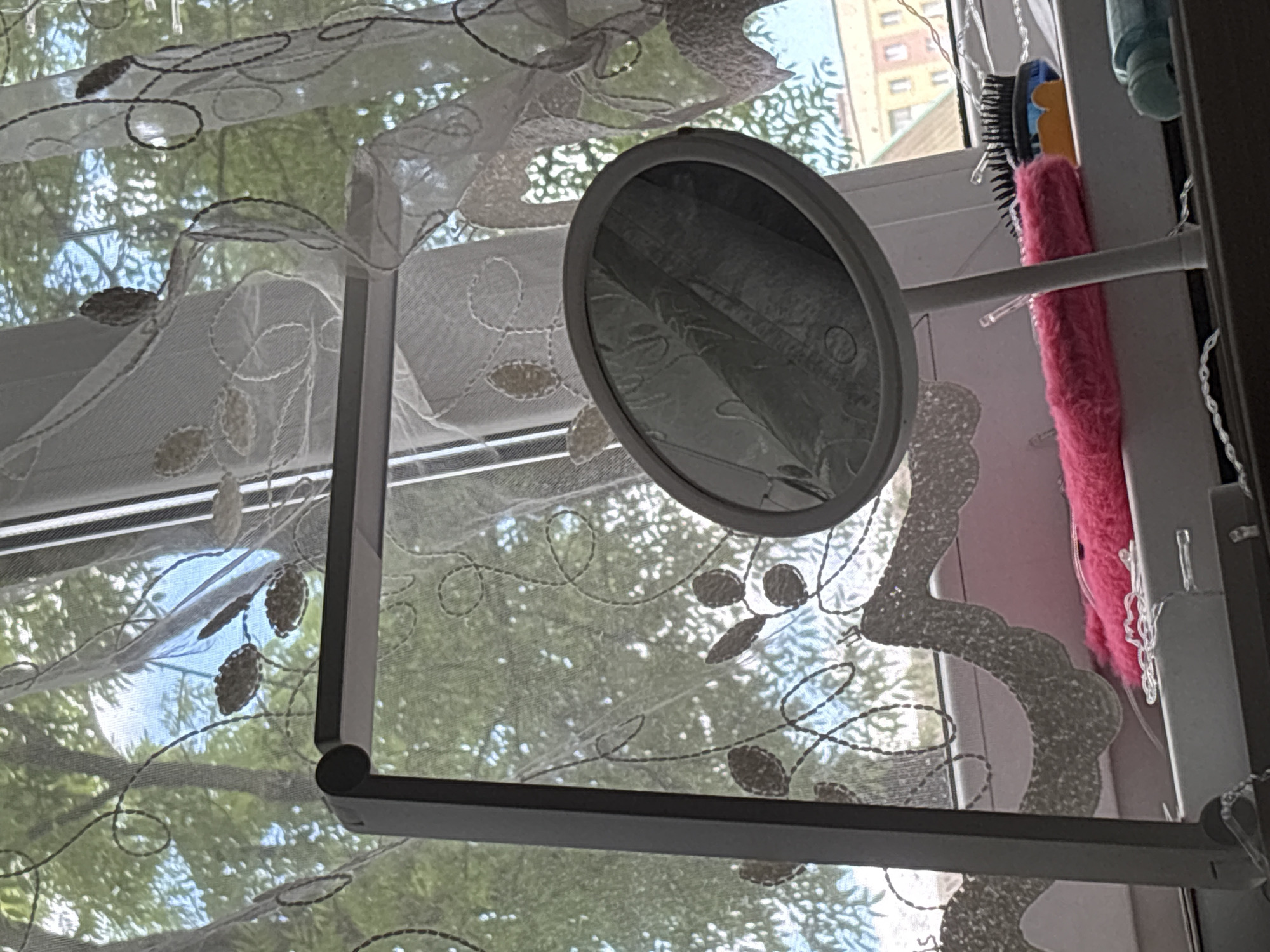 The image shows a close-up view of a window sill in an apartment in Minsk, Belarus. On the sill, there is a modern desk lamp, an oval hand mirror, a pink fluffy object, and a hairbrush with a blue and yellow handle. A sheer curtain with embroidered floral patterns hangs in front of the window, obscuring much of the view outside. Through the curtain, trees and the upper floors of buildings are visible, suggesting an urban setting. The overall impression is one of a domestic interior, possibly a vanity area near a window.