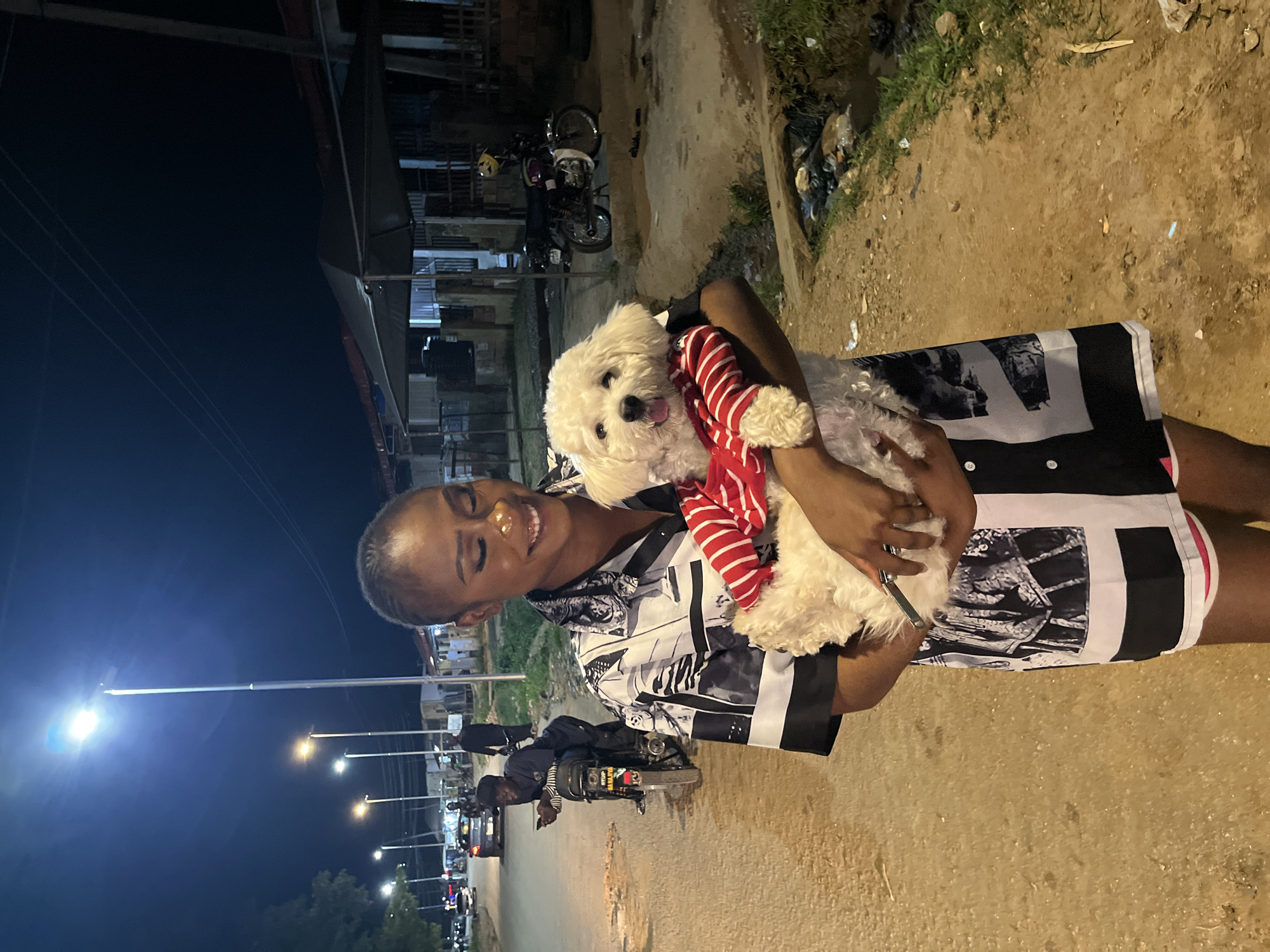 A Black woman with a shaved head and striking makeup smiles as she holds a small white dog wearing a red and white striped shirt. She is wearing a black and white patterned button-up shirt dress and pink shorts. The setting is outdoors at night, illuminated by streetlights. In the background, a dirt road is visible, with motorcycles parked on the side and a few people walking or standing. Buildings and tents line the street. The environment appears to be a bustling urban area, likely within Abuja, Nigeria, suggested by the presence of motorcycles, street vendors, and the overall atmosphere of a developing city at night.