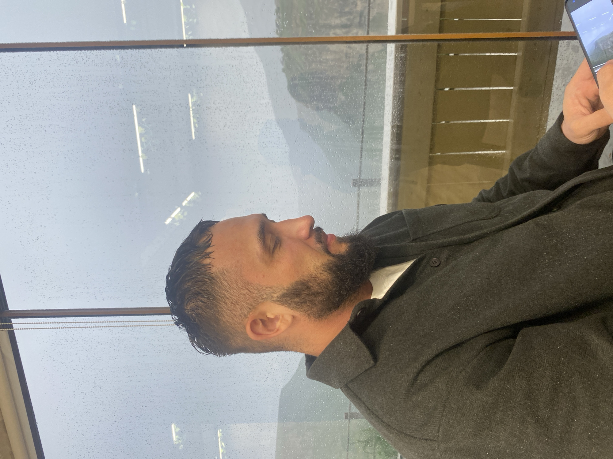 A man with a beard and a dark grey shirt is sitting by a window overlooking a misty mountain landscape. The window is covered in raindrops, suggesting it is raining outside. The man is looking down at a smartphone in his hands. The background shows blurred mountains and trees. The scene is likely in a cafe or restaurant in Khunzakh, Russia, a town known for its scenic mountain views.