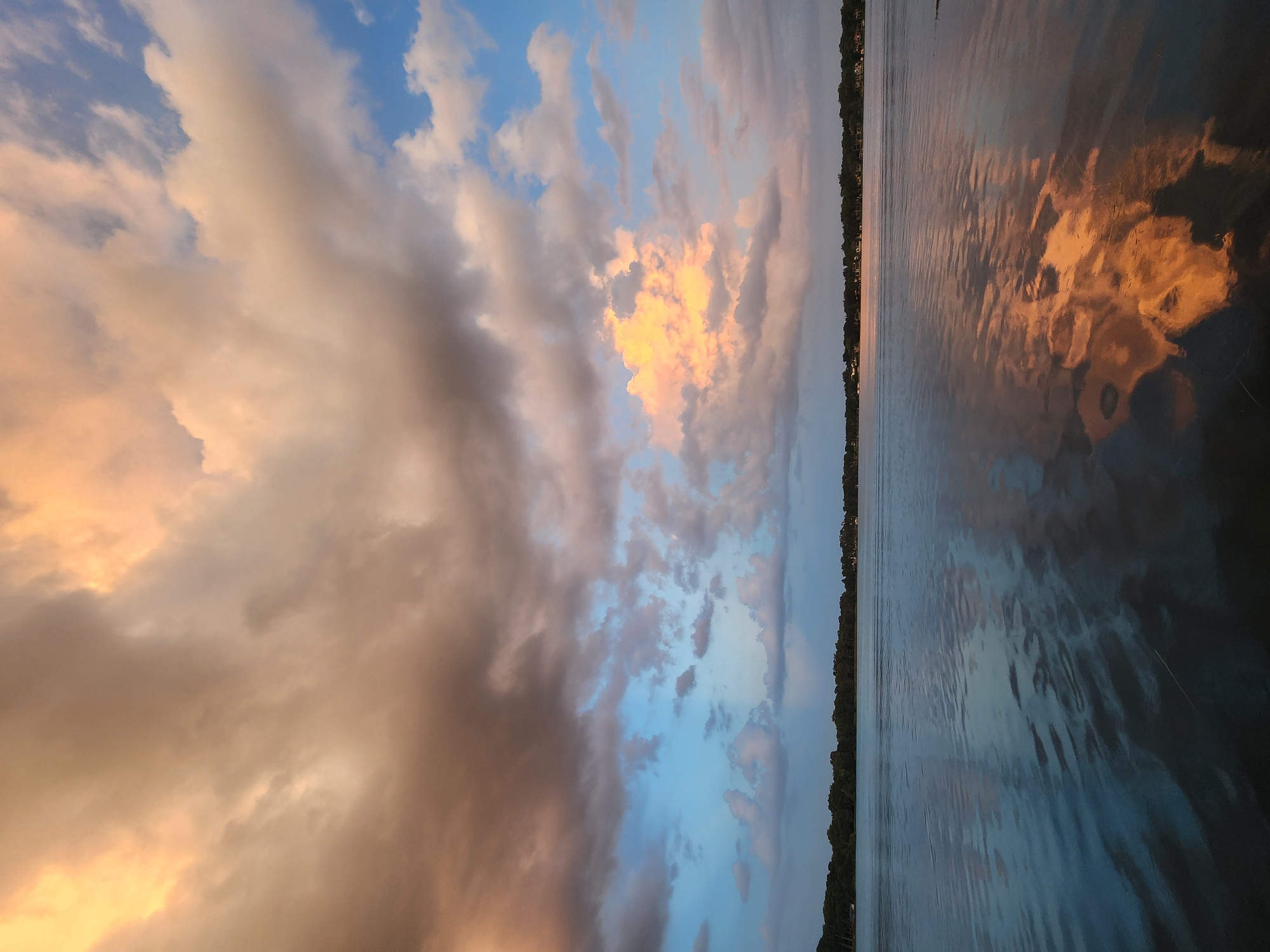 This is a photograph of a body of water, likely a lake, on a partly cloudy day. The sky is filled with dramatic clouds, illuminated by the setting sun with hues of orange, pink, and yellow. The water reflects the sky and the distant tree-lined shore, creating a mirror-like effect. The far bank of the water is lined with a dense strip of trees, behind which are visible houses and structures, suggesting a residential area. The water is calm with gentle ripples, and there are no people or boats visible in the scene. The overall impression is one of serene natural beauty and the tranquility of a late afternoon or early evening. This scene could be found at a location such as Lake Virginia or another of the many lakes within Winter Park, Florida, known for its picturesque lakes and abundant natural beauty.
