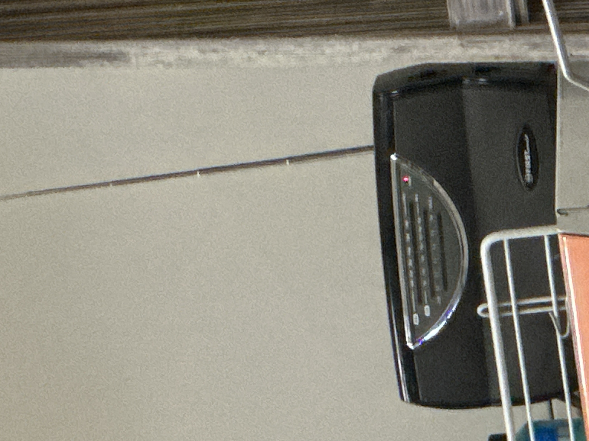 An indoor, close-up shot captures a collection of objects against a light gray wall and a textured, darker ceiling or upper structure. The central focus is a black electronic device, likely a speaker or radio, positioned vertically on the right side of the frame. It features a transparent or translucent curved front grille with several horizontal lines and a small, circular red indicator light illuminated on the upper left side of the grille. A vertical logo, possibly "SHERWOOD" or "SHER," is faintly visible on the right side of the device.
To the left of the device, a thin, horizontal metal rod or wire stretches across the wall, supported by a few small white attachments. On the far right, partially visible, are white metal bars forming part of a rack or shelf system. An orange or copper-colored object is tucked within this white metal frame. The background wall is plain and uniform in texture, while the ceiling area above shows horizontal dark lines, suggesting corrugated material or paneling. There is no discernible activity or event taking place, and no people are present. The lighting suggests an interior setting, with no specific time of day or weather indicated. The overall scene appears utilitarian, potentially a storage area, garage, or workshop in Pleven, Bulgaria.