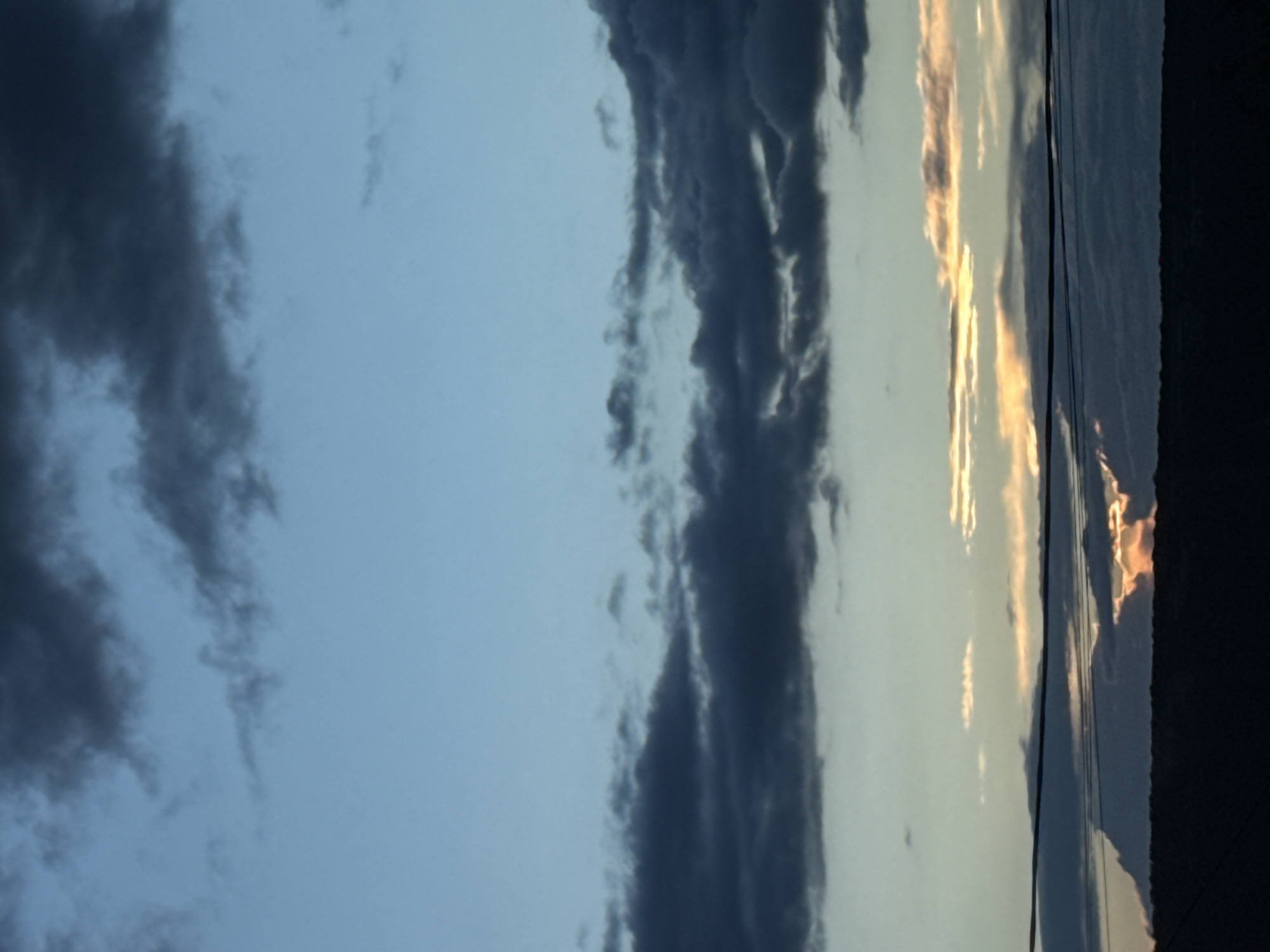 The image shows a twilight or sunset sky over Перовское сельское поселение, Ukraine. Dark, striated clouds dominate the left portion of the frame, contrasting with a lighter, illuminated section to the right, where golden light is visible. Power lines are strung across the lower-right, and a dark object (possibly a building) is visible at the extreme right edge.