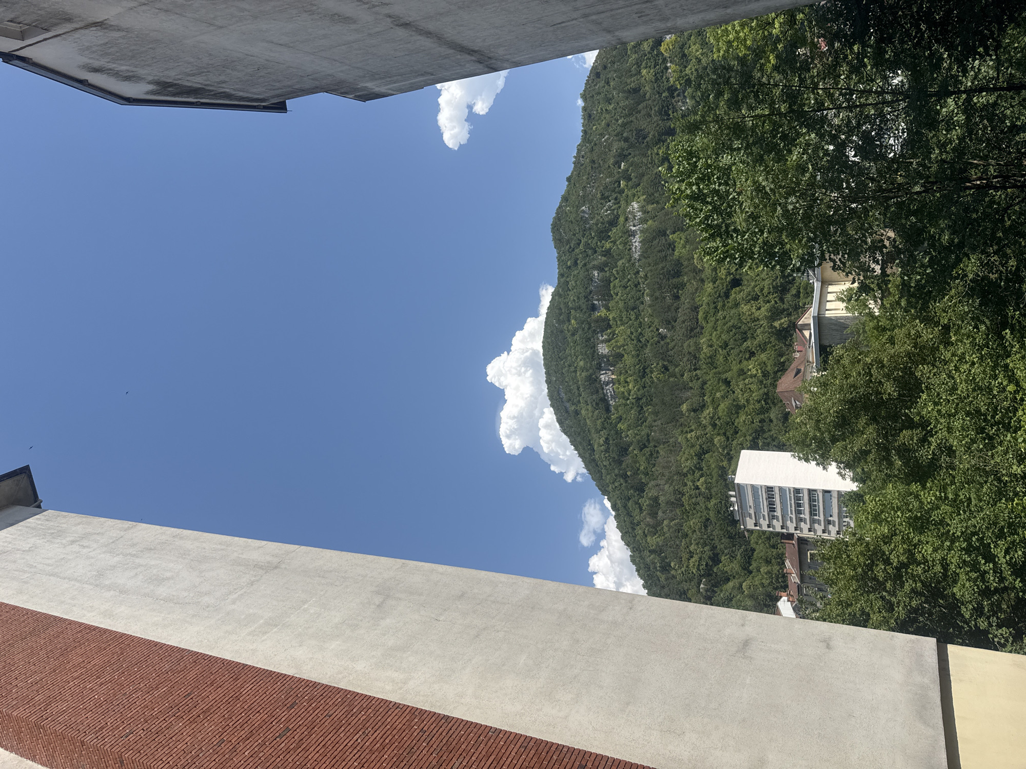 The image depicts a view from a window or balcony in Saint-Claude, France, looking out towards a lush, green mountain under a bright blue sky with scattered white clouds. In the foreground, two building facades frame the view: a reddish-brown brick wall on the left, angled and extending upwards, and a gray concrete wall on the right, also angled and extending upwards. Between these structures, a cluster of trees with dense green foliage obscures the lower portion of the scene. In the middle ground, several buildings are visible, including a tall, white, multi-story apartment building with numerous windows, and other residential structures with red-tiled roofs. The mountain in the background is densely covered with trees, with some rocky outcrops visible near its peak. The overall impression is a serene, sunlit urban landscape bordering a natural environment.