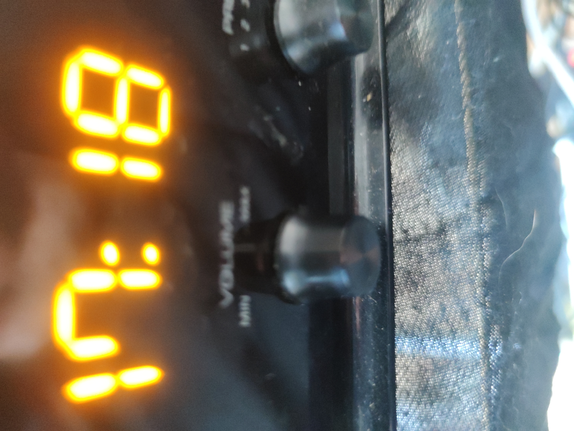The image shows a close-up of a clock radio. The digital display indicates the time as 01:18. There is a volume control knob below and a tuning control knob above. A textured surface, potentially cloth, is to the right of the radio. The location is unknown.