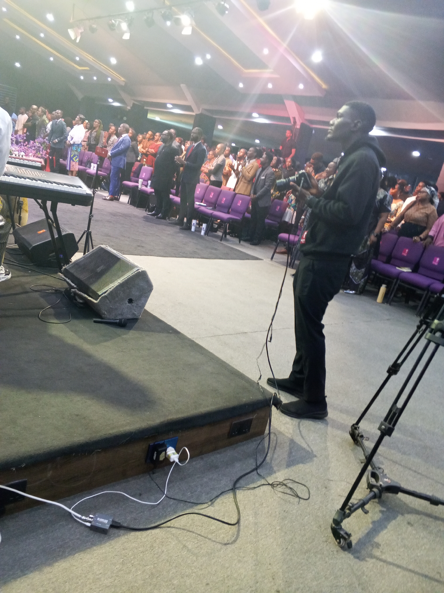 The image shows the interior of a large room in Abuja, Nigeria, likely a church or auditorium. A crowd of people, mostly adults, is standing in the background. Some are dressed in suits and formal attire. A man in black clothing stands in the foreground, operating a camera. A keyboard, speakers, and a camera tripod are near him. Power cords are plugged into an outlet on the edge of the platform. The lighting is bright, with many overhead lights illuminating the space. Purple chairs are visible behind the crowd.