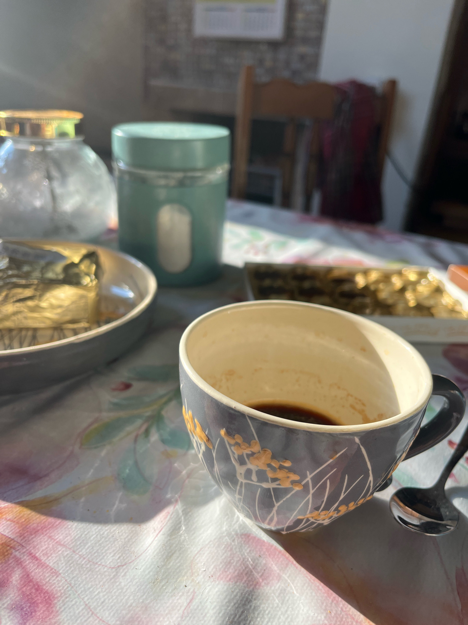 A close-up, low-angle shot captures a breakfast scene on a table in Cheshnegirovo, Bulgaria. The focal point is a dark blue ceramic mug with a floral design, partially filled with dark liquid, likely coffee or tea. Next to the mug lies a silver spoon. In the background, a plate holds a wrapped item, possibly food, covered in gold foil. To the left, a clear glass jar with a golden lid and a teal canister are visible. The table is covered with a light-colored tablecloth featuring a floral pattern. Sunlight streams into the room, casting shadows and highlighting the objects. The environment suggests an indoor setting, possibly a dining area.