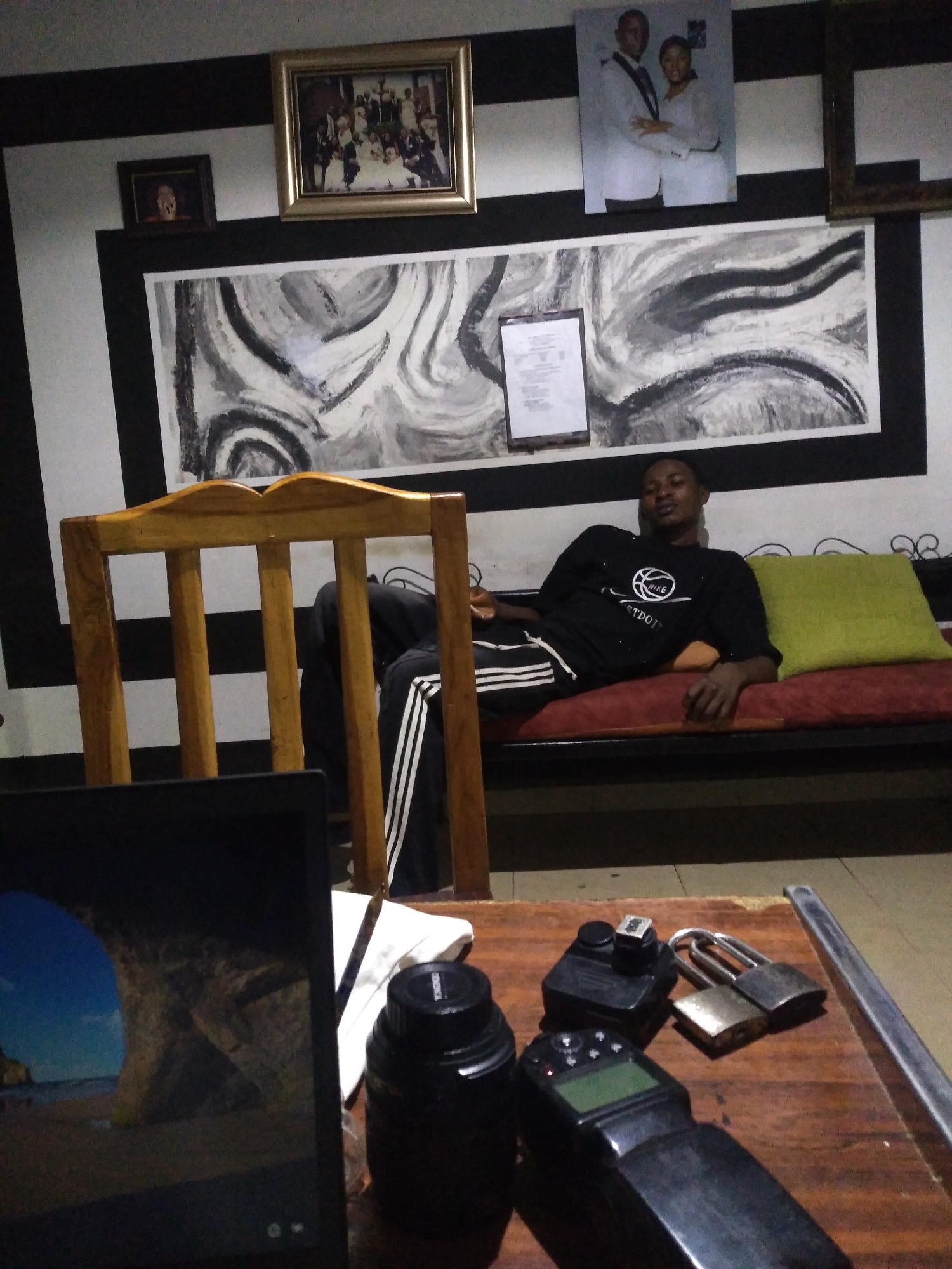 In an indoor setting, a young Black man is reclining on a sofa. He is wearing a black t-shirt with a white Nike logo and black shorts with white stripes. The sofa is red with a green cushion. In front of the sofa, a wooden chair is positioned, and beyond that, a laptop displays a scenic screensaver. On a wooden table in the foreground, several objects are visible: a camera lens, a flash unit, and two padlocks. The wall behind the sofa is decorated with a large abstract painting in black and white, and several framed photographs are hung above it. The room has black and white striped walls.