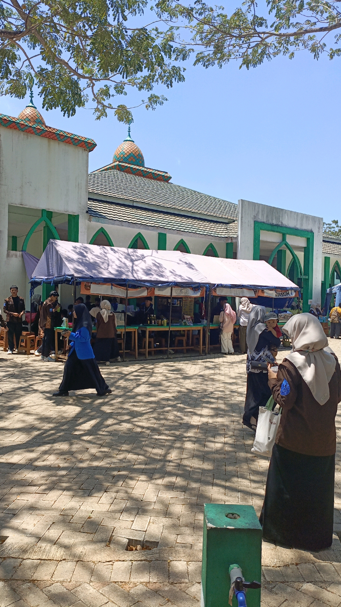 The image depicts an outdoor market scene in front of a mosque in Desa Pabahanan, Pelaihari, Tanah Laut, Indonesia. The mosque features green and orange tiled domes and white walls with green arched accents. A large white tent is set up in front of the mosque, displaying various goods on tables. Several individuals, mostly women wearing hijabs, are browsing or working at the stalls. Some people are walking on a paved courtyard, with dappled sunlight creating shadows on the ground from overhead trees. In the foreground, a green trash receptacle with a faucet is visible.