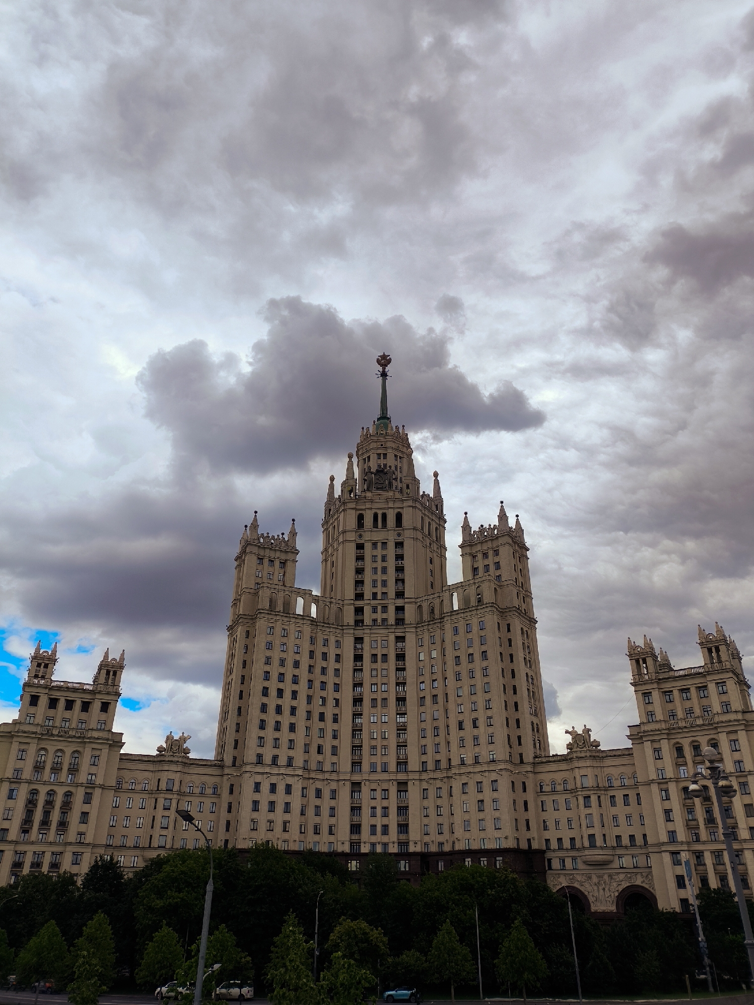 The image shows a low-angle view of a grand, Stalinist-era skyscraper against a cloudy sky. The building is made of light-colored stone and features multiple towers, spires, and intricate architectural details. Rows of windows punctuate the facade of the massive structure. In the foreground, a dense line of dark green trees creates a stark contrast with the pale building and the dramatic sky. A street with a few cars and a lamppost is visible at the bottom of the frame, hinting at an urban setting. The overall impression is one of imposing architecture under a brooding sky.
