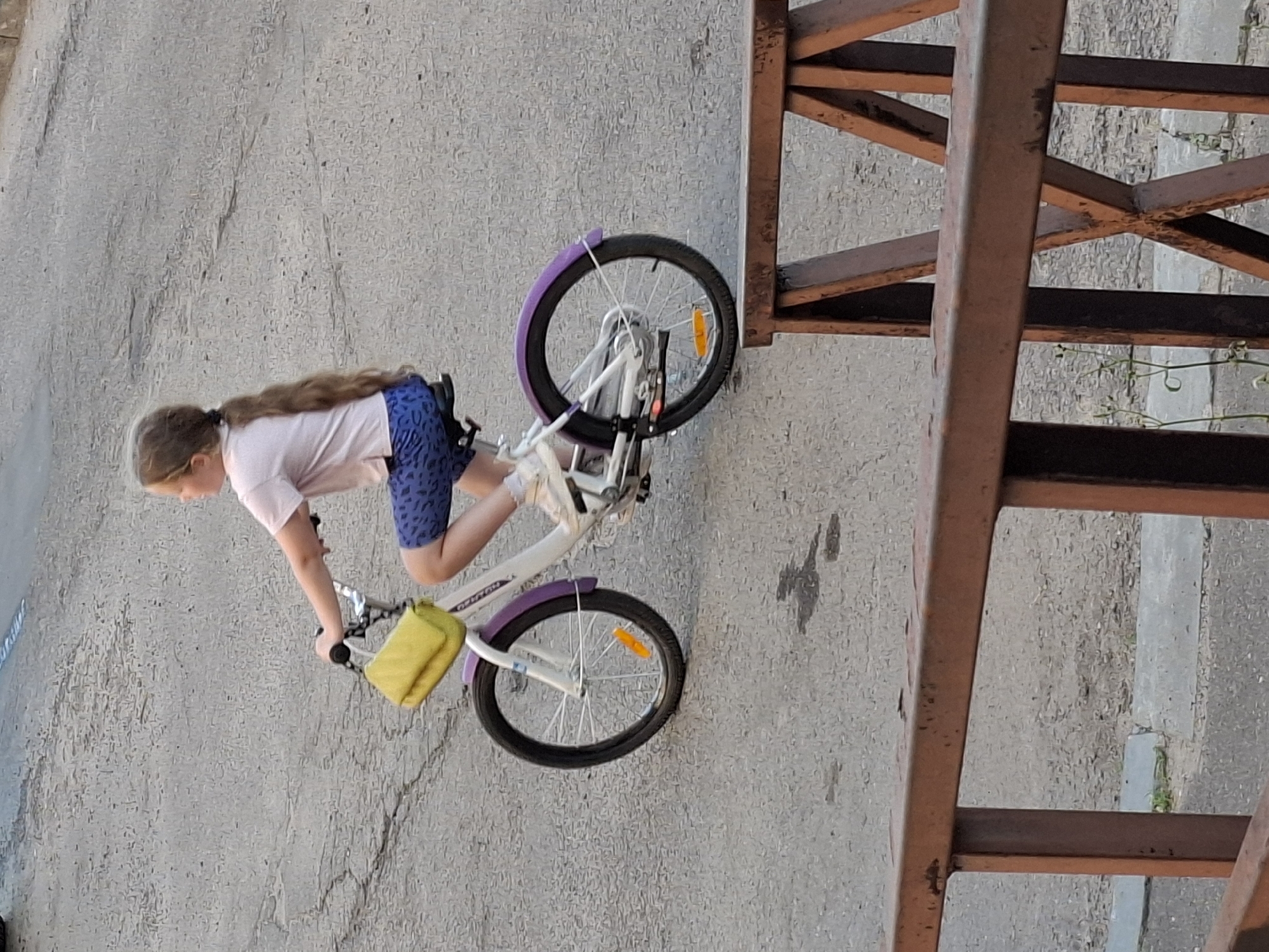 An overhead shot captures a young girl with long, brown hair braided down her back riding a white bicycle with purple fenders. She wears a light pink t-shirt and blue patterned shorts. A yellow pouch is attached to the handlebars. The bicycle has a silver frame and black tires. The girl is positioned on a gray asphalt surface, possibly a street or sidewalk, with some cracks visible. In the foreground, a rusty metal railing or fence partially obstructs the view. The background shows more of the asphalt and a hint of greenery.