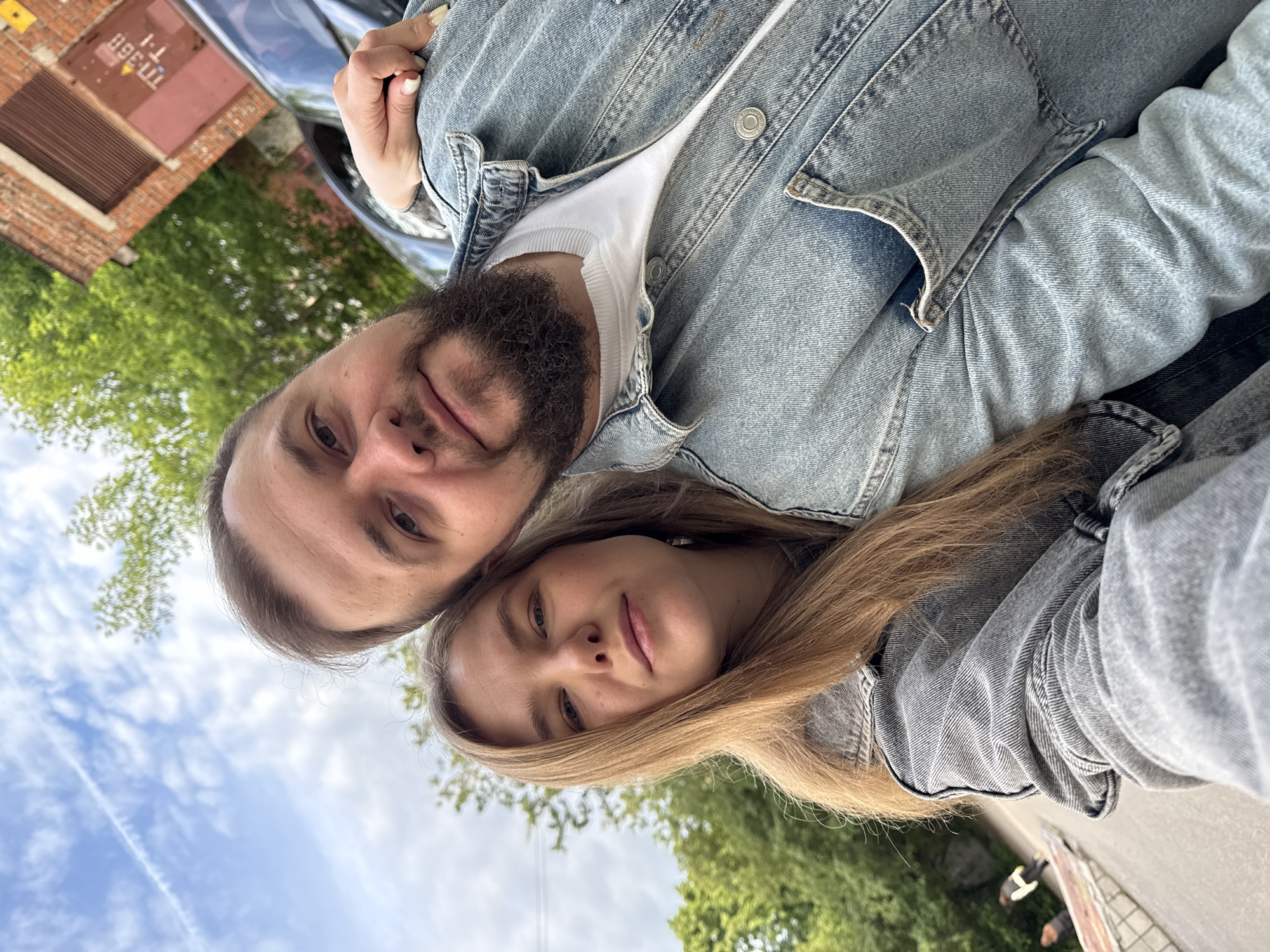 A couple is taking a selfie outdoors. The man has dark hair, a beard, and is wearing a denim jacket over a white shirt. The woman has long blonde hair and is also wearing a denim jacket. They are close together, seemingly embracing. Behind them are trees and a blue sky with clouds. In the background is a building made of red brick. The location is in the city of Vladimir, Russia.