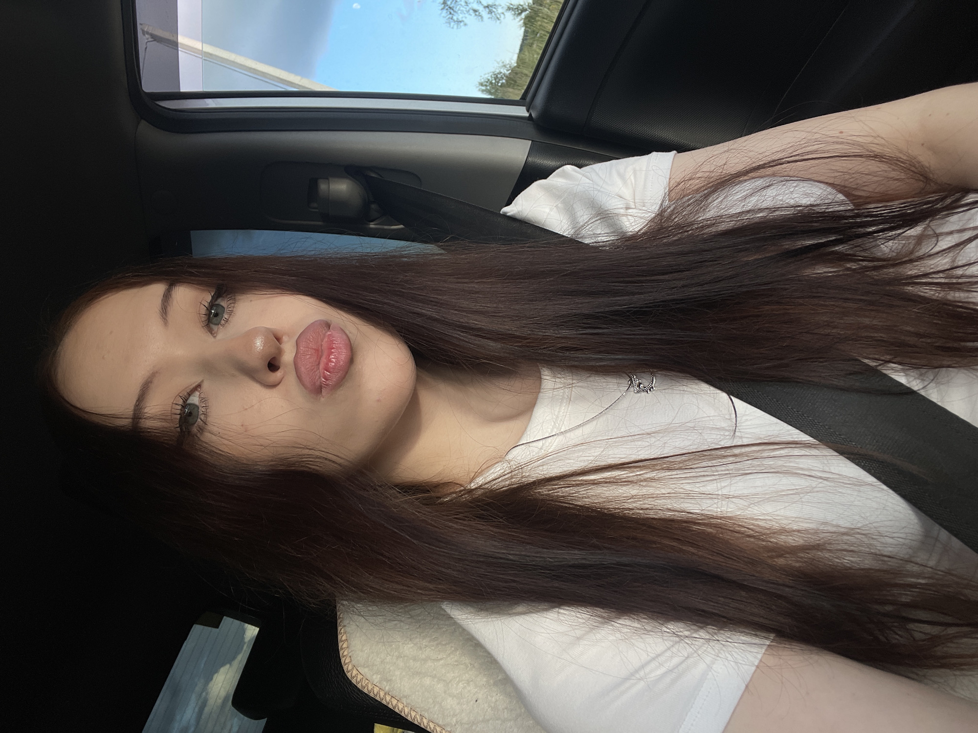The media file is a selfie of a young woman taken inside a car, likely in Gavrilovka, Russia. She has long, dark brown hair that falls over her shoulders and chest. She is wearing a white t-shirt and a silver necklace. Her makeup is visible, with emphasis on her eyes and lips, which are slightly parted. The image is taken at an angle, showing a portion of the car's interior, including a seatbelt and the roof lining. Outside the car, the sky is visible through the sunroof and window, appearing to be daytime with some trees and foliage in the background. The overall mood appears casual and relaxed, possibly during a car journey. There is no visible text in the media file.
