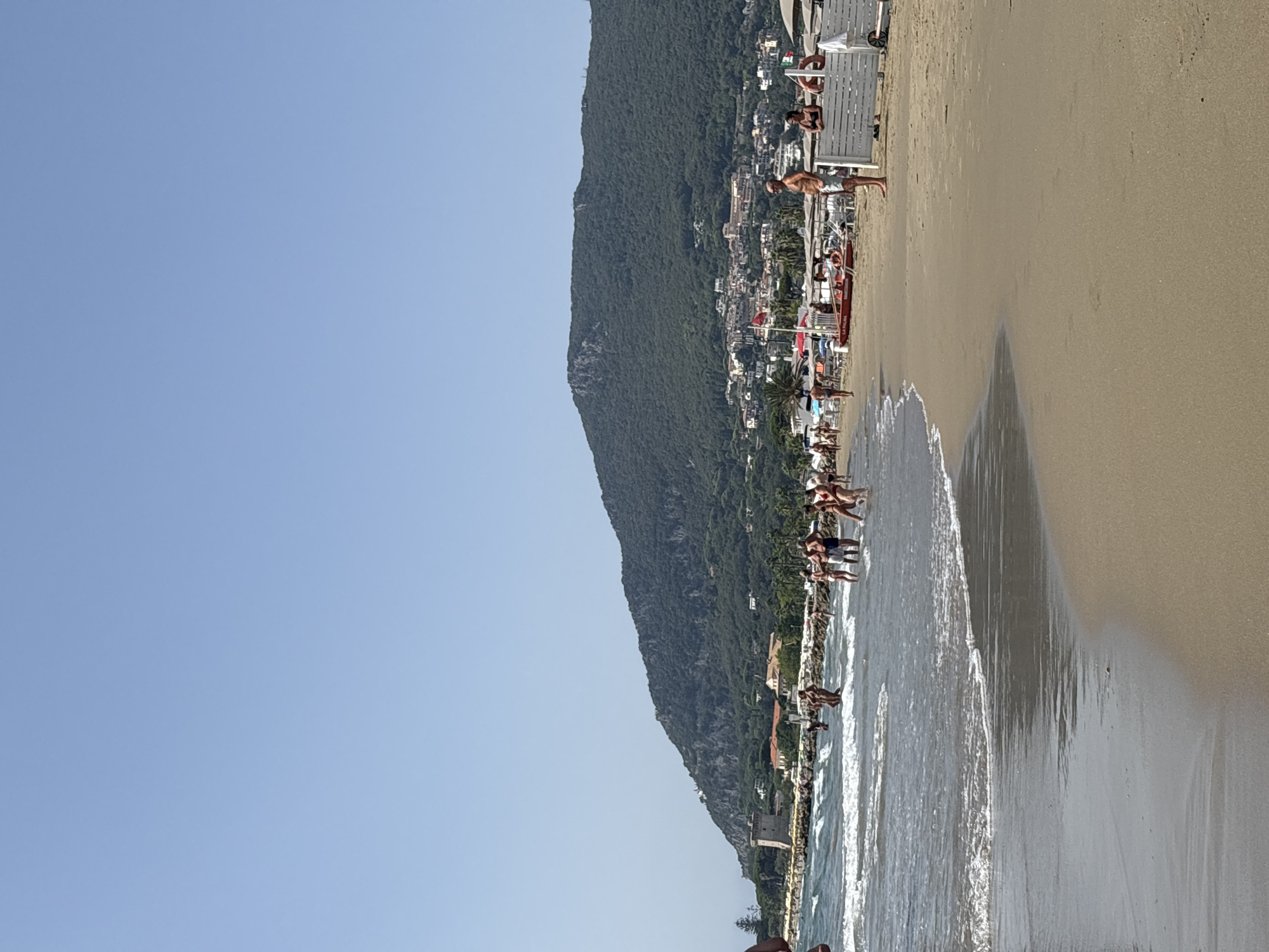 A bright, sunny day at a beach in San Felice Circeo, Italy. The image captures a panoramic view of a sandy coastline with a calm, blue sea. In the foreground, gentle waves lap onto the shore. A group of people are visible in the shallow water, some wading and others standing around. Further back, a line of beach umbrellas and loungers suggests a well-equipped beach resort. Behind the beach, a lush, green mountain dominates the skyline, its slopes dotted with buildings forming a coastal town. A historical tower, possibly part of an ancient fortification, stands on the left side of the image, near the water's edge. The sky is clear and a vibrant blue, characteristic of a summer day.