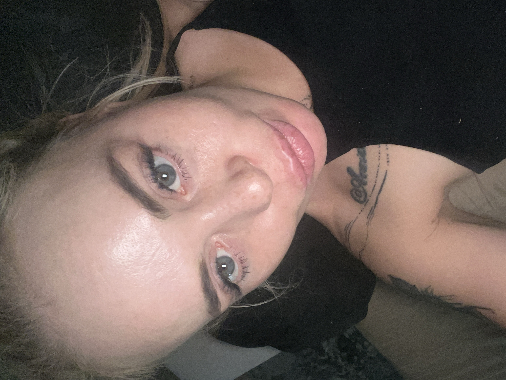 A close-up, angled shot shows a person lying down, looking towards the camera. Their eyes are open, and their lips are slightly parted. The person has blonde hair and appears to be wearing makeup, with noticeable eyeliner and mascara. There are tattoos visible on their arm and shoulder area. The background is dark, suggesting the image may have been taken in low light conditions. The overall mood is intimate and casual. There is no discernible text in the image.