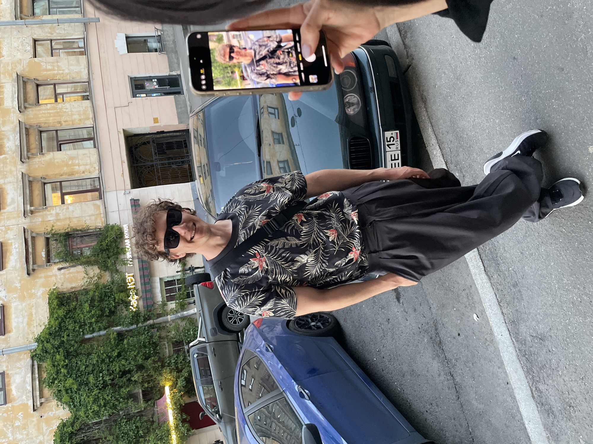 The image captures an outdoor street scene in Saint Petersburg, Russia. A male subject is positioned slightly left of center, facing forward and smiling. He has curly brown hair, wears black sunglasses, a dark short-sleeved shirt with a tropical pattern of green leaves and red/orange flowers, dark pleated trousers, and dark sneakers with white soles (a Nike swoosh is visible on the right shoe). A black cross-body bag is worn over his left shoulder. His left hand is in his pocket.
On the far right, the right arm and hand of a second individual are visible, holding a horizontally oriented smartphone (an iPhone) with light-colored manicured fingernails. The phone's screen displays a live preview of the male subject.
The background consists of a multi-story building with a weathered, yellowish-beige facade. Green ivy heavily covers portions of the building, particularly around windows. A neon yellow sign in Cyrillic, "Кафе КЭТ" (Cafe KET), with "ГРУЗИНСКАЯ КУХНЯ" (Georgian Cuisine) below it, is visible on the facade, partially obscured by ivy. A striped awning extends below the cafe sign. To the right of the main building, a section with a light pink/peach facade features a dark entrance door and a window. An ornate black metal gate is situated between building sections.
Three vehicles are parked on the asphalt street. Immediately behind the male subject is a dark green BMW E39 5-Series sedan, displaying a Russian license plate "EE 15. RUS" on its front bumper. To the left, a blue Hyundai Accent sedan with black multi-spoke wheels is parked. Further in the background, a dark green pickup truck with a spare tire mounted vertically on its bed is visible. The street surface includes a white painted line. The scene is illuminated by natural daylight.