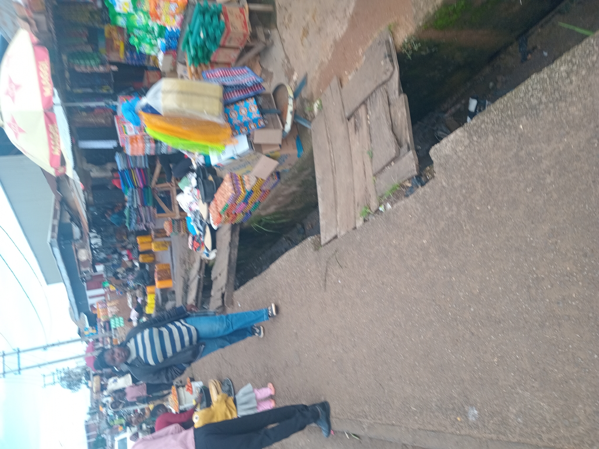 The image depicts a street scene in Bukuru, Nigeria, likely a market area. A woman in a striped jacket and blue jeans walks on a paved sidewalk, facing away from the camera. To her left, a small child in a yellow top and grey skirt with pink leggings stands next to a person wearing black pants and a pink top. In the background, various market stalls are visible with goods displayed, including bags of products, stacked items, and a prominent red and white Maggi umbrella. A narrow, open drain runs alongside the sidewalk, spanned by wooden planks in some sections. The environment appears to be outdoors, with buildings and overhead wires suggesting an urban setting.