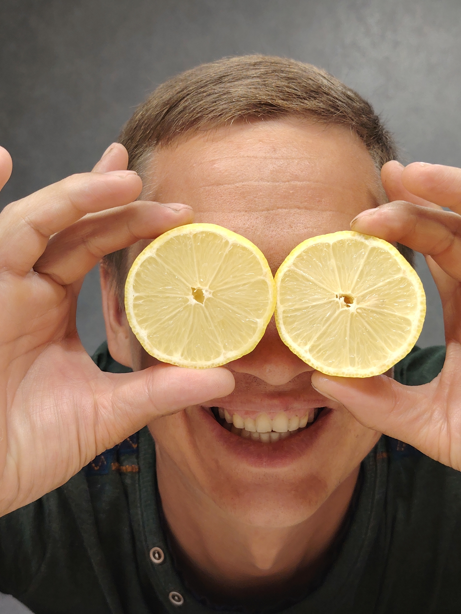 A close-up, chest-up, medium shot features a Caucasian male with short brown hair and light skin, positioned centrally. He is holding two lemon slices up to his eyes, with his fingers obscuring parts of his face and the lemons. The man is smiling, revealing his teeth. He is wearing a dark green t-shirt with visible buttons. The background is a plain, dark grey wall. No identifiable location in Chaikovsky, Russia, is depicted in this image.