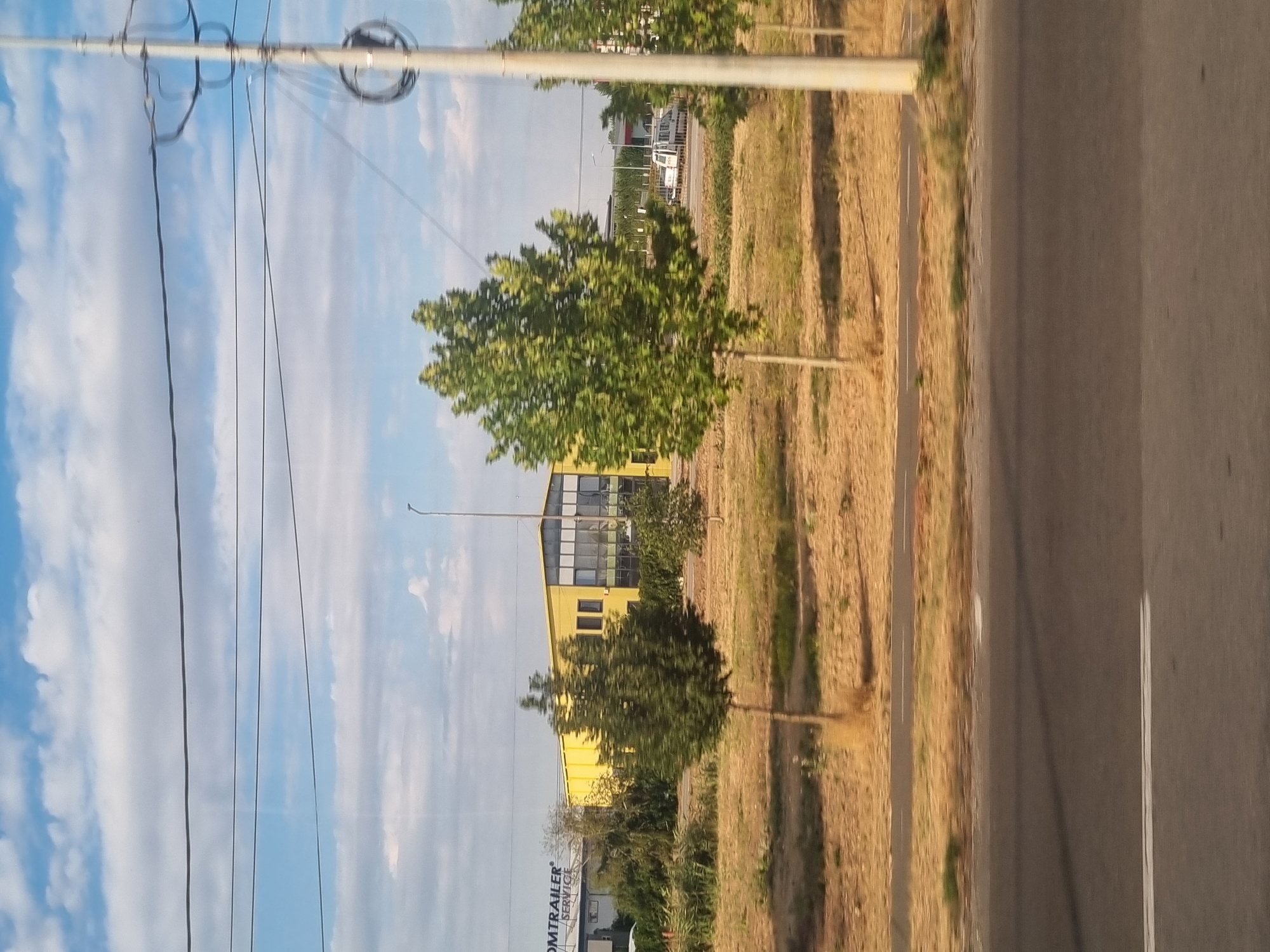 The image depicts a suburban or industrial area in Sântion, Romania, on a cloudy day. In the foreground, there is a paved road with white lane markings. Beyond the road, a dry, grassy field stretches towards a line of trees and a yellow building. The building appears to be an industrial or commercial facility, with large windows and a sign that reads "OMTRAILER SERVICE." Several trees are scattered throughout the landscape, with one prominent, slender tree in the middle ground. In the background, more trees and buildings are visible, along with utility poles and power lines crisscrossing the sky. A white van or minibus is parked on the far side of the road. The overall impression is one of a functional, possibly light industrial or commercial zone on the outskirts of a town.