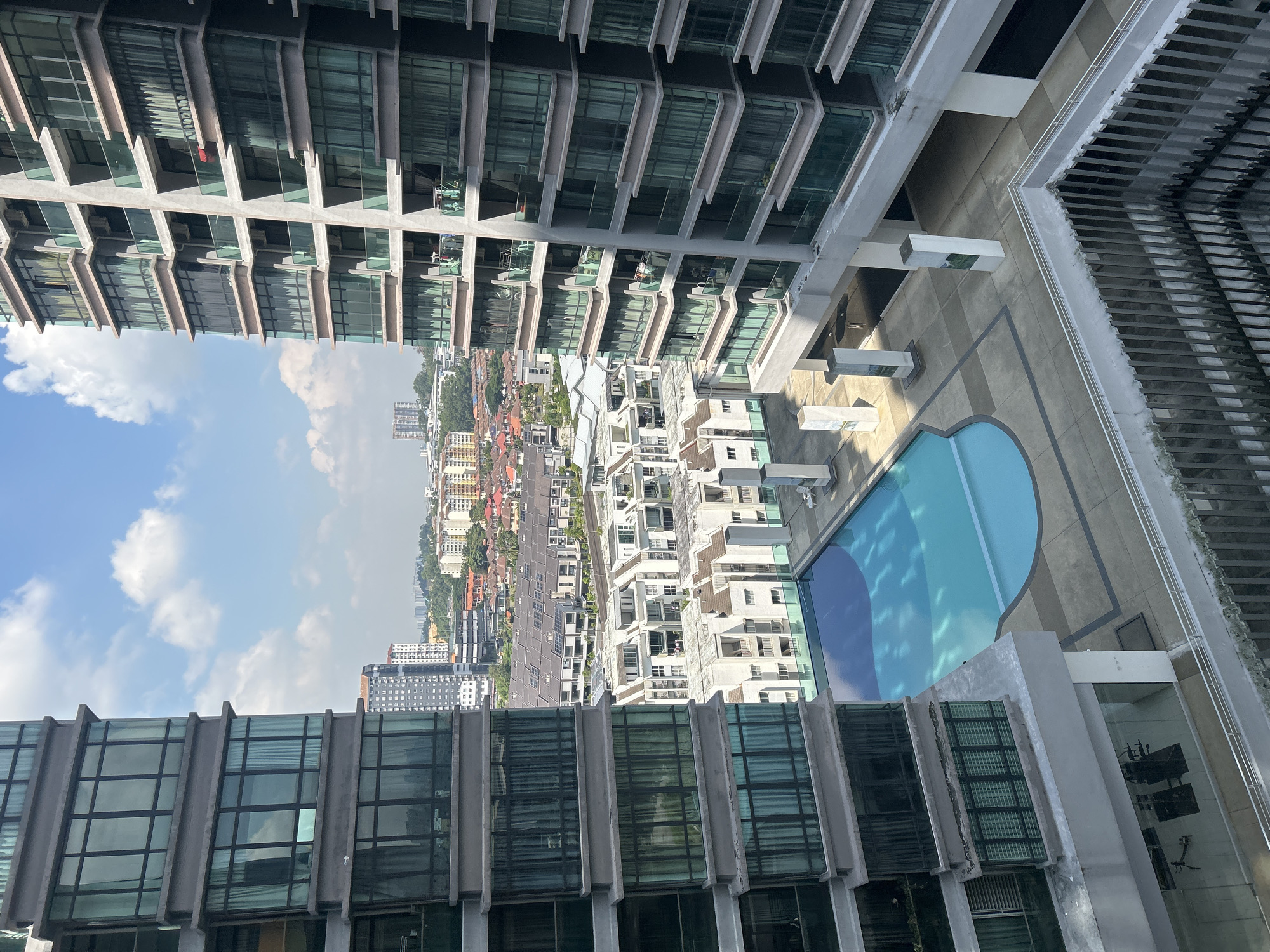 The image shows an elevated view of a cityscape, likely taken from a high-rise building. In the foreground, there are sections of two similar high-rise buildings with rows of glass windows and concrete supports. Between the buildings is an open area that features a swimming pool, with its water displaying varied shades of blue.
Beyond this, a lower-rise urban area is visible, with buildings of varying heights and architectural styles, including residential houses with red roofs, interspersed with greenery. In the distance, more high-rise buildings punctuate the skyline, suggesting the presence of a city center.
The sky occupies a significant portion of the upper left area, exhibiting a gradient of light to medium blue, dotted with scattered white clouds.
Based on architectural styles and urban density, this is likely Kuala Lumpur, Malaysia.