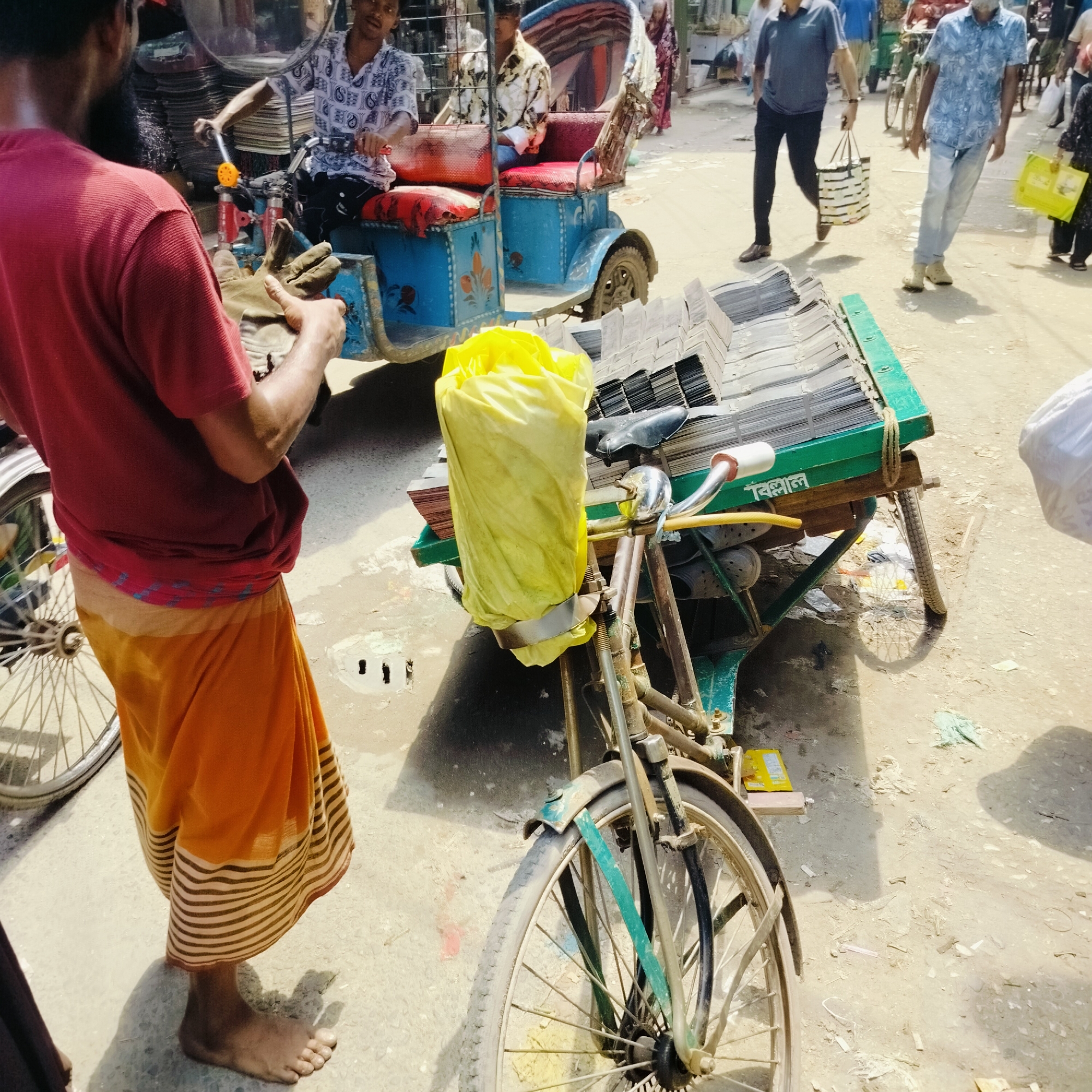 The image captures a bustling street scene in Dhaka, Bangladesh. A bare-footed man wearing a red shirt and orange and white striped sarong stands next to a bicycle modified into a vendor's cart. The cart is loaded with corrugated metal sheets and a yellow plastic bag. The cart has "বিলাল" painted on the front frame. In the background, a blue auto-rickshaw with two passengers is visible, along with pedestrians walking by. One man in a blue shirt and black pants is carrying a striped bag. The setting appears to be a marketplace or commercial area with a dirt ground.