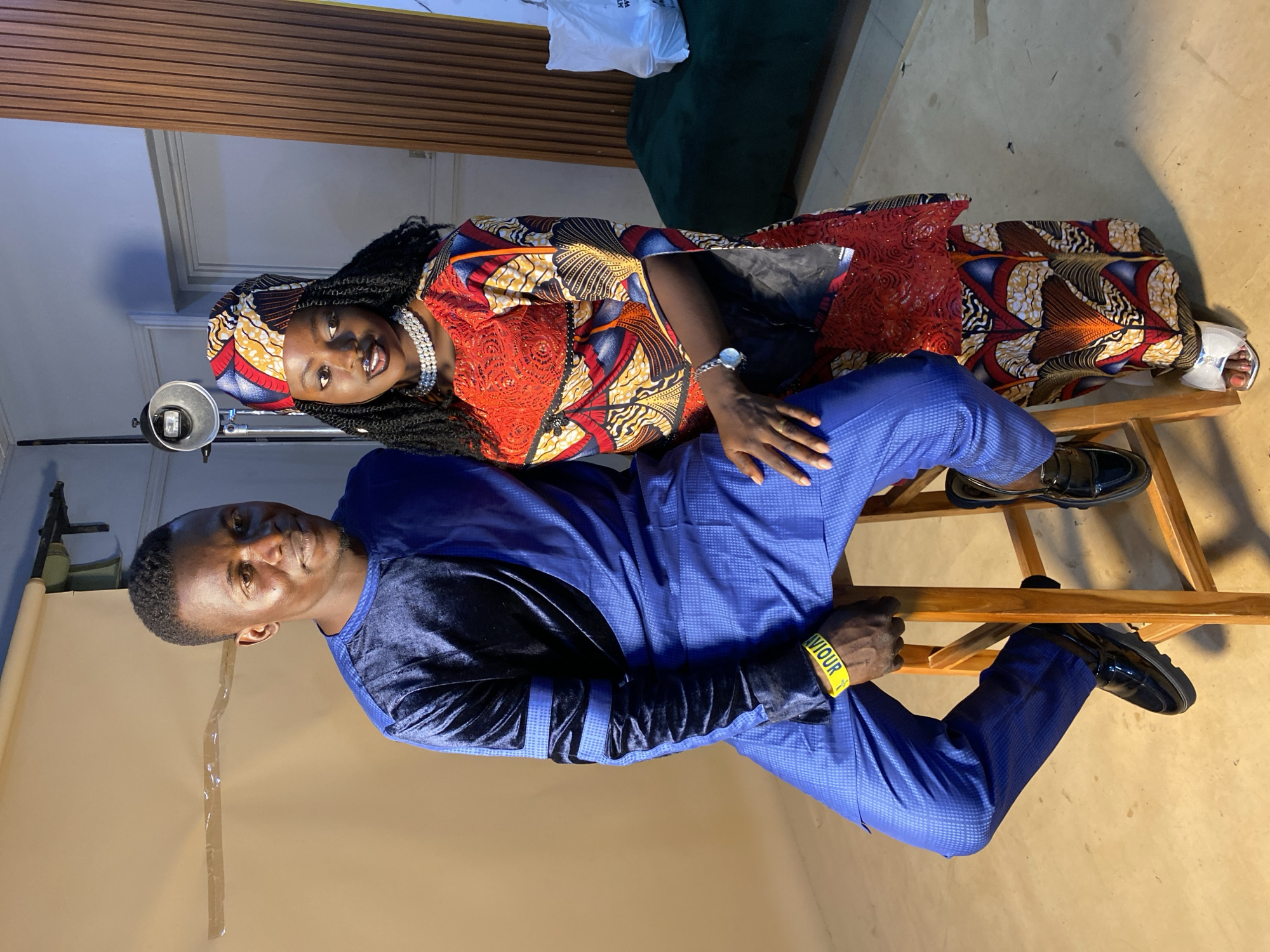 A man and woman pose together in what appears to be a photography studio in Bukuru, Nigeria. The man is seated on a wooden chair, wearing a blue outfit and shiny black shoes. He has a yellow wristband with black writing on it. The woman stands behind him, wearing a vibrant, patterned dress with red details and a matching headscarf. She also wears a beaded necklace. She places her hand on the man’s knee. In the background is a beige backdrop, a lighting stand, and a green couch.