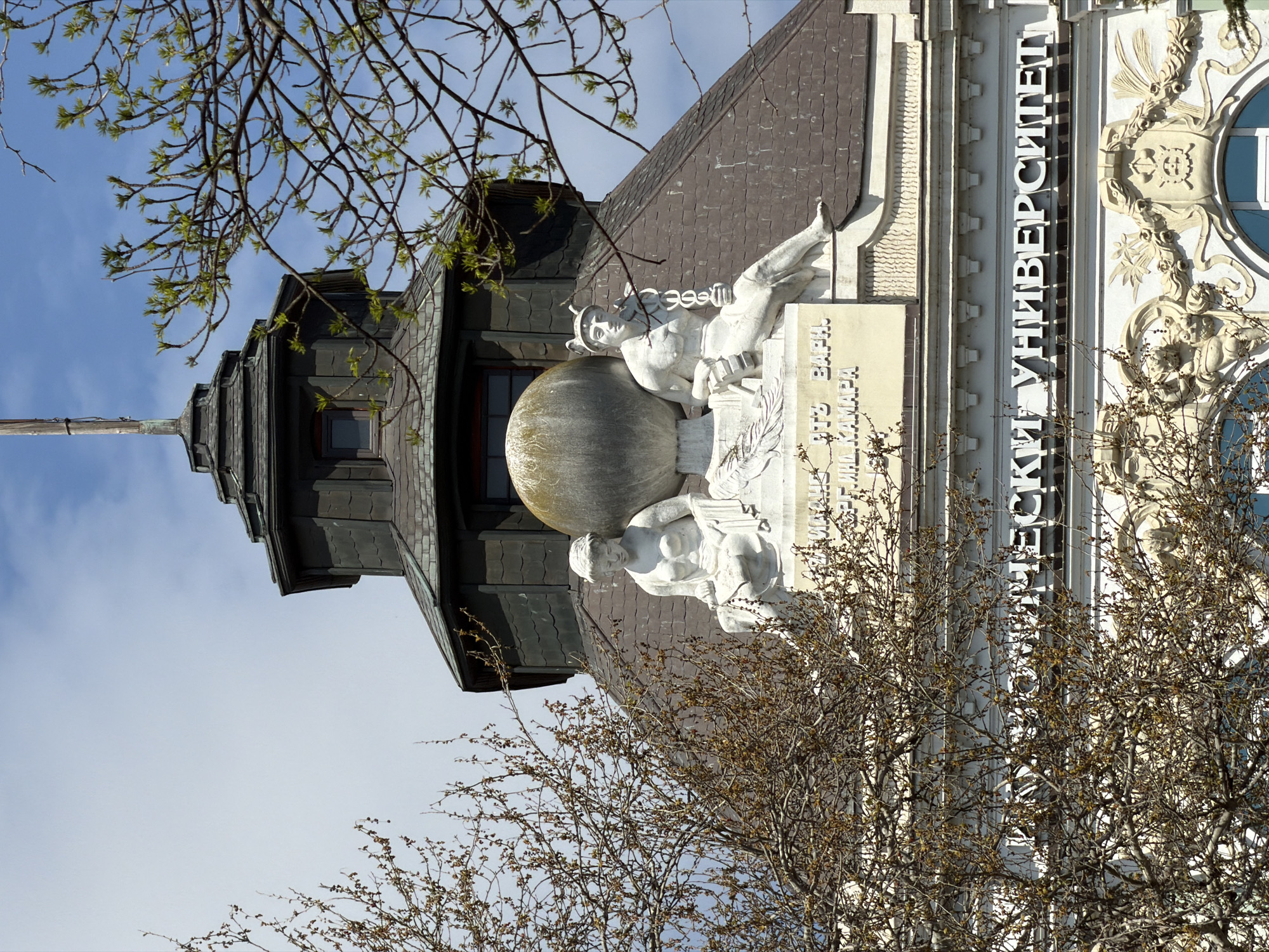 An architectural shot captures a section of a university building in Varna, Bulgaria, under a bright, clear blue sky. The image is oriented vertically (rotated 90 degrees counter-clockwise), showing the building's facade and roofline.
The building features a dark, multi-faceted roof made of small, possibly slate or metal, tiles. A slender, dark-colored spire-like structure with small windows rises from the left side of the roof. The main facade, appearing to be light-colored stone or concrete, is richly decorated.
Prominently displayed are large, white, classical-style sculptures. One group features two figures: an upper figure, possibly a scholar or deity, wearing a helmet and looking upward, with a book or tablet in hand; below this, another figure supports a large, textured spherical object, likely a globe or celestial sphere. Below these sculptures, Cyrillic text is partially visible, reading "ВАРНА" (Varna) and "ИМ. ХРАБАР" (named after Hrabar), pointing to the Varna Free University "Chernorizets Hrabar."
Further to the right on the facade, large white Cyrillic letters clearly spell out "УНИВЕРСИТЕТ" (UNIVERSITET), confirming the building's function as a university. Ornate decorative carvings, including stylized foliage and a crest-like emblem, adorn the facade around the lettering.
In the foreground, bare tree branches with emerging light green leaves, indicative of early spring, partially obscure the view of the building and its details. Denser, darker branches also appear lower down, creating more visual obstruction. The overall impression is one of academic grandeur, set against a pleasant, sunny day.