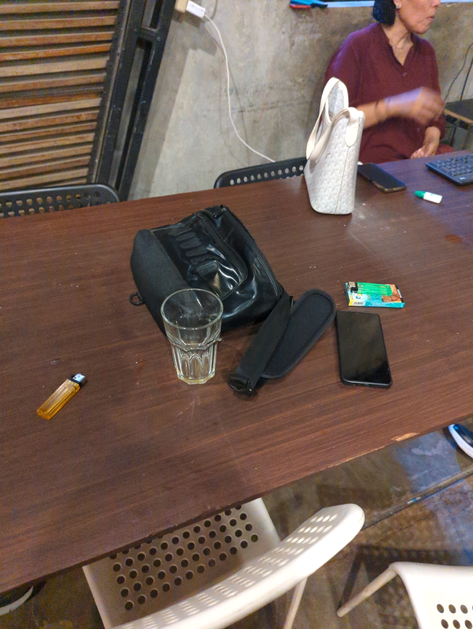 A medium shot shows a wooden table with various items on it, as well as a person in the background. In the foreground, an orange lighter lies to the left of a clear glass with a faceted design. Next to the glass is a black bag with a shiny surface and a strap. To the right of the bag is a pack of gum. A black smartphone is placed below the gum pack. In the background, a person wearing a maroon shirt sits with their right arm extended, possibly interacting with something on the table. A white tote bag rests on the table near the person. The environment appears to be indoors, with a concrete wall and wooden blinds visible. There are also white chairs with perforated surfaces.