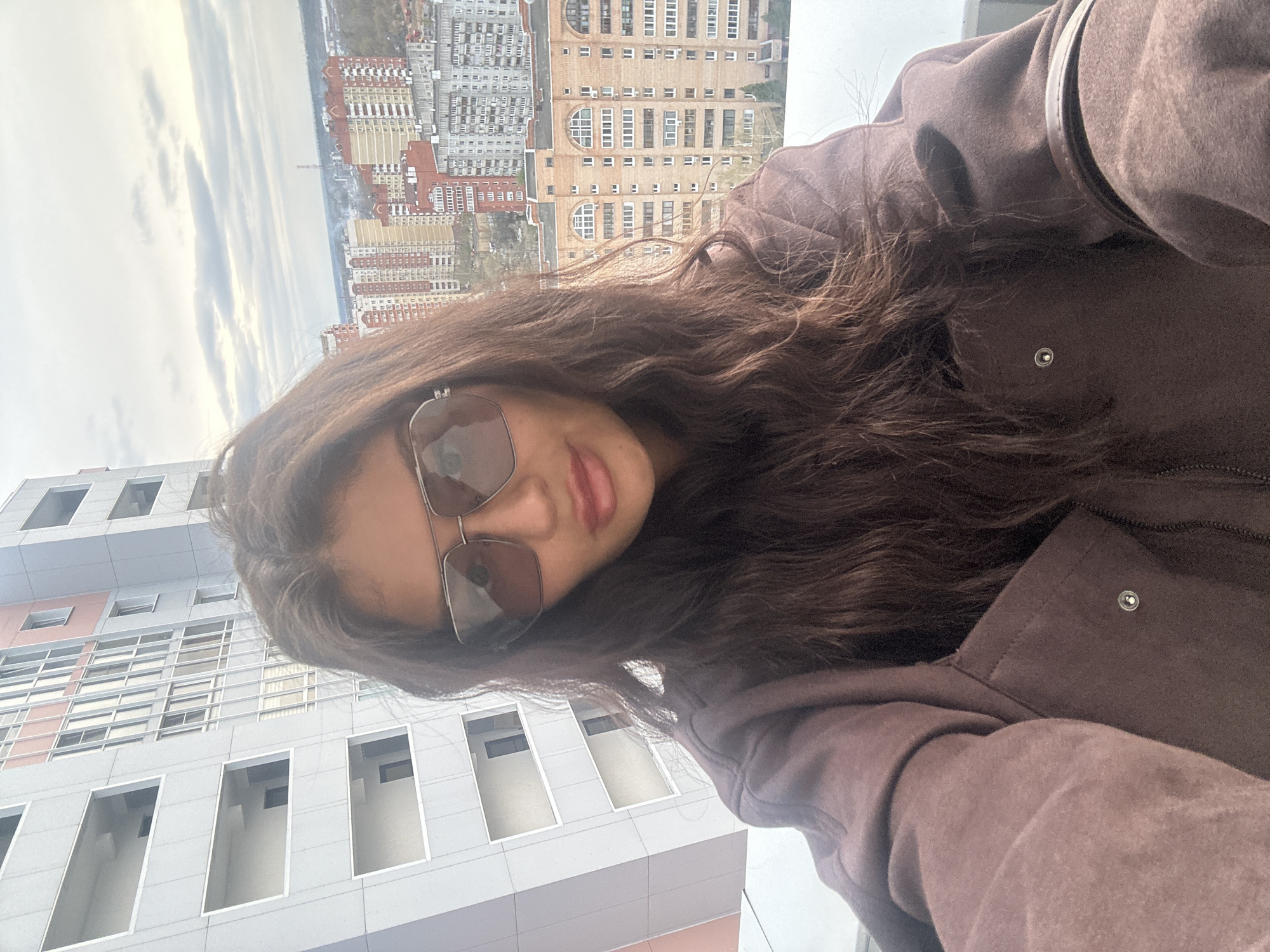 A medium shot depicts a woman posing outdoors, angled to include architectural features and a cityscape in the background. The woman is wearing sunglasses and a brown jacket. The architectural elements suggest she is standing near a high-rise building with a modern design, characterized by rectangular windows and a predominantly gray color scheme. The cityscape in the distance shows a collection of buildings varying in height and design, with a river or large body of water visible in the background under a cloudy sky. Based on the architectural style and urban layout, the location is identified as Krasnogorsk, Russia.