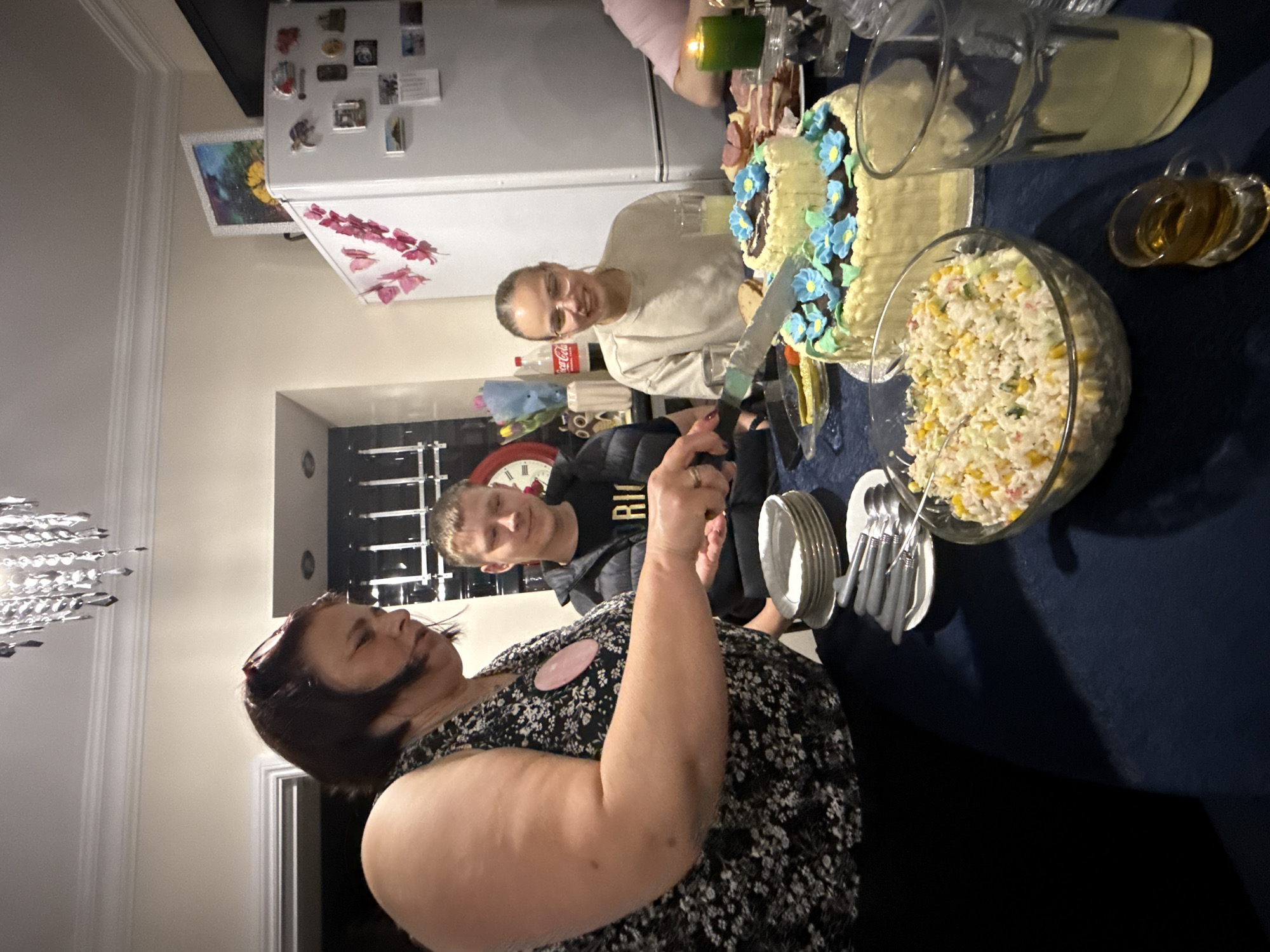 The image captures an indoor gathering, likely a celebration, with several people around a table laden with food. The foreground features a woman in a floral dress with a pink badge, her arm raised. To her right, a young boy in a black jacket and a "RICH" t-shirt looks on. Behind them, a woman with glasses cuts a cake decorated with blue flowers. The table is covered with a dark blue cloth and holds plates, cutlery, a large bowl of salad, a cake, and a pitcher of a light-colored liquid. In the background, a white refrigerator adorned with magnets and a clock on the wall can be seen. A chandelier hangs from the ceiling in the lower left corner. The location is not specified, nor any indication of the city of Great Yarmouth, United Kingdom.