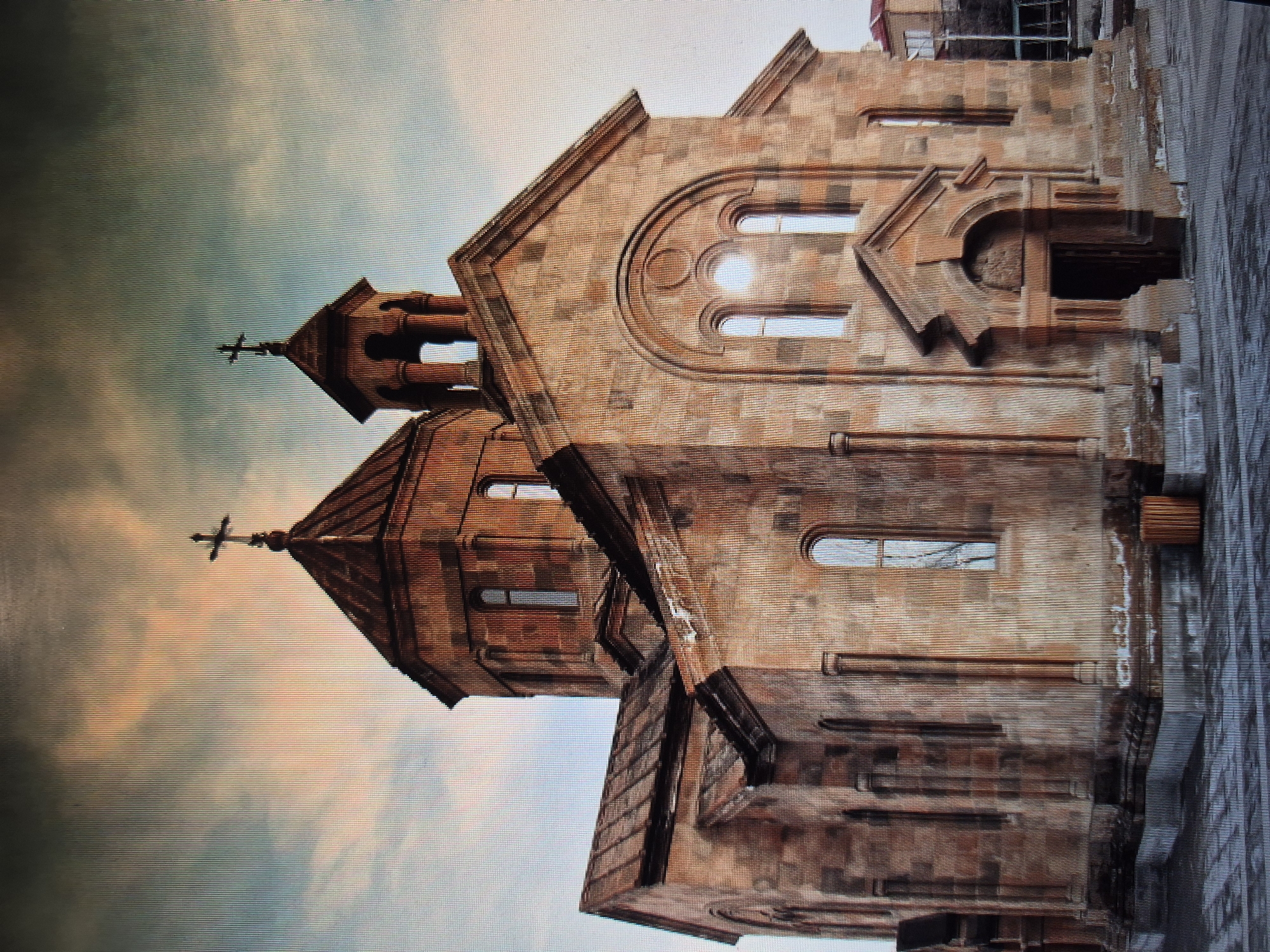 The image shows the Saint Anna Church in Yerevan, Armenia. The stone facade of the church is visible, featuring arched windows, a central entrance, and decorative elements. The church has two pointed domes, each topped with a cross. The sky behind the church is a mix of pale blues and pinks, possibly indicating dawn or dusk. A building with scaffolding can be seen partially in the background on the right side of the image. A grey paved surface is visible on the right side of the image. The photo appears to be taken at a slight angle from below.