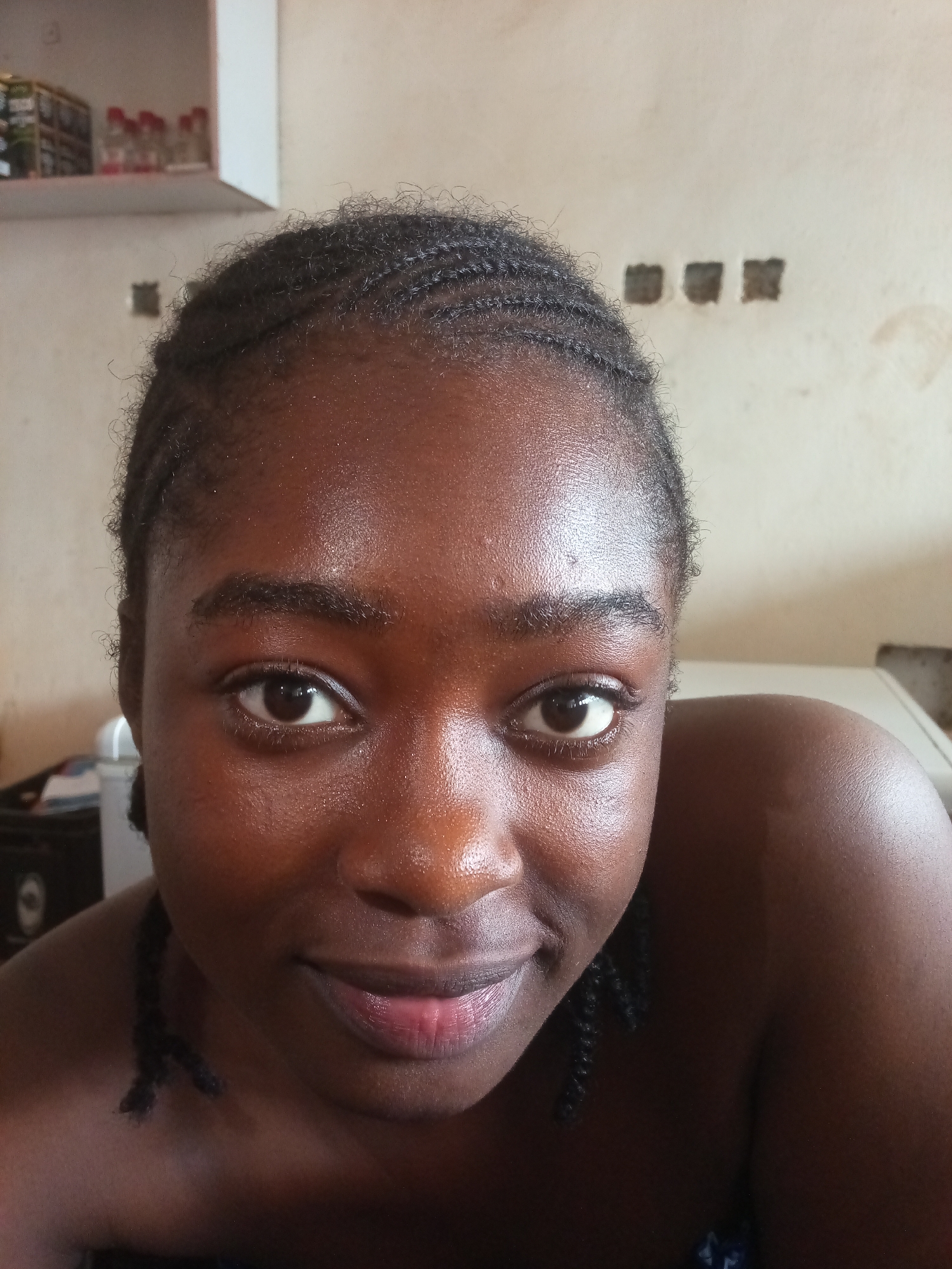 A close-up shot, taken indoors, focuses on the face of a young Black woman with braided hair and dark skin. She is looking directly at the camera with a subtle smile. Her eyes are brown and her lips are tinted pink. The background is out of focus, showing a beige wall with some unidentifiable objects and a shelf with items on it. A portion of her shoulder and upper chest are visible. The lighting is warm and highlights the natural sheen of her skin. There is no indication of the city of New Stadium, Nigeria, in the image itself, and no objects or context suggest this specific location.
