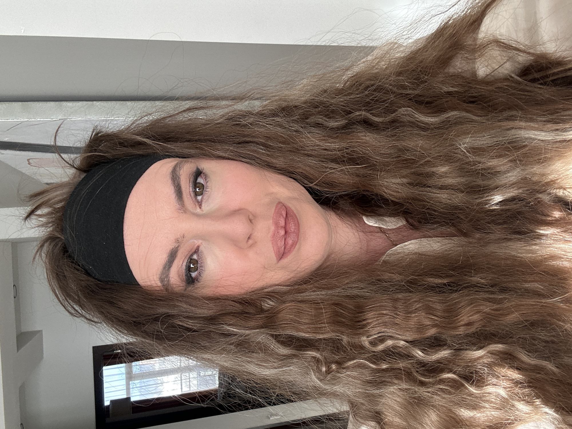 A woman with long, wavy, light brown hair is centered in the image. She is wearing a black headband. Her facial features include fair skin, brown eyes, and full lips. She appears to be indoors. Through a window in the background, a window with blinds is visible. It's not possible to identify if this picture was taken in Tbilisi, Georgia.