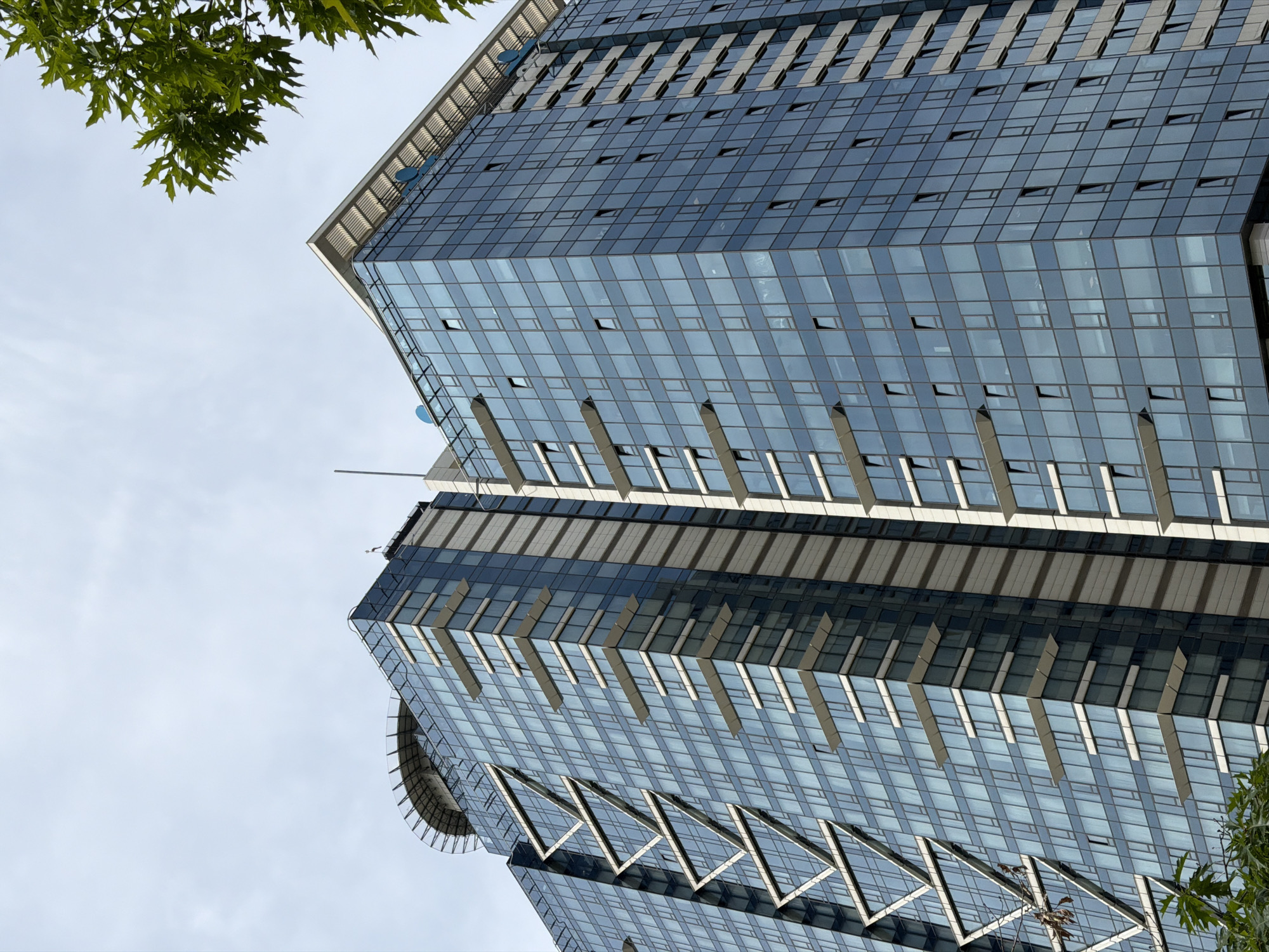 The image shows a low-angle view of a modern glass-clad building in Sofia, Bulgaria. The building features a grid-like pattern of windows and is segmented into distinct sections with projecting horizontal elements. It appears to be a multi-story structure with a helipad visible on one section. The sky is partially visible in the background, along with some foliage from trees in the foreground, which suggests the building is situated in an urban environment with some greenery.