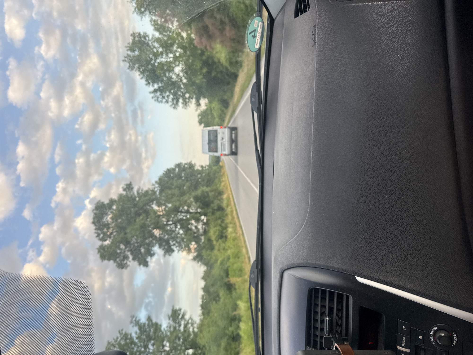 The image was taken from inside a vehicle driving on a rural road, likely near Draganovtsi, Bulgaria. The sky is partly cloudy with a mix of blue sky and white clouds. Lush green trees and vegetation line both sides of the asphalt road. In the distance, a white van is driving ahead on the same road. The foreground shows the interior of the vehicle, including the dashboard and windshield wipers. A circular sticker with green and white markings is visible on the lower right side of the windshield.