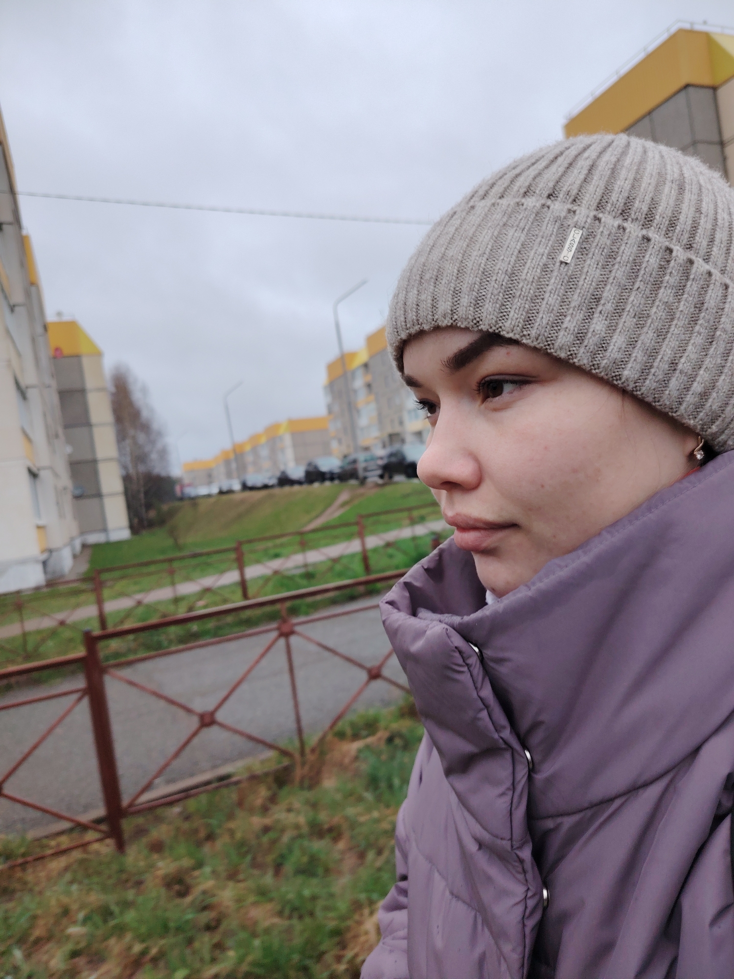 A candid, waist-up portrait features a young woman outdoors, looking towards the right with a pensive or neutral expression. She has fair skin, dark eyebrows, and brown eyes. She is wearing a light grey ribbed knit beanie, which has a small metallic tag on the front, possibly inscribed with the word "LUXURY." Her outerwear is a purple or plum-colored puffer jacket with a high collar and silver snap buttons. A small, light-colored earring is visible in her right ear.
The setting appears to be a residential area in Markovskii, Russia, under an overcast sky, suggesting cool weather. In the foreground, a reddish-brown metal fence with a geometric, crisscross pattern is visible. Beyond the fence, a grassy slope leads to a street with several parked cars. The background is dominated by multi-story residential buildings, likely apartment blocks, characterized by their light-colored facades and distinct bright yellow upper sections or roofs, a common architectural style in some Russian cities. Bare trees are scattered in the mid-ground, further indicating autumn or early winter. The scene depicts an ordinary outdoor moment, possibly a walk or a pause during the day.