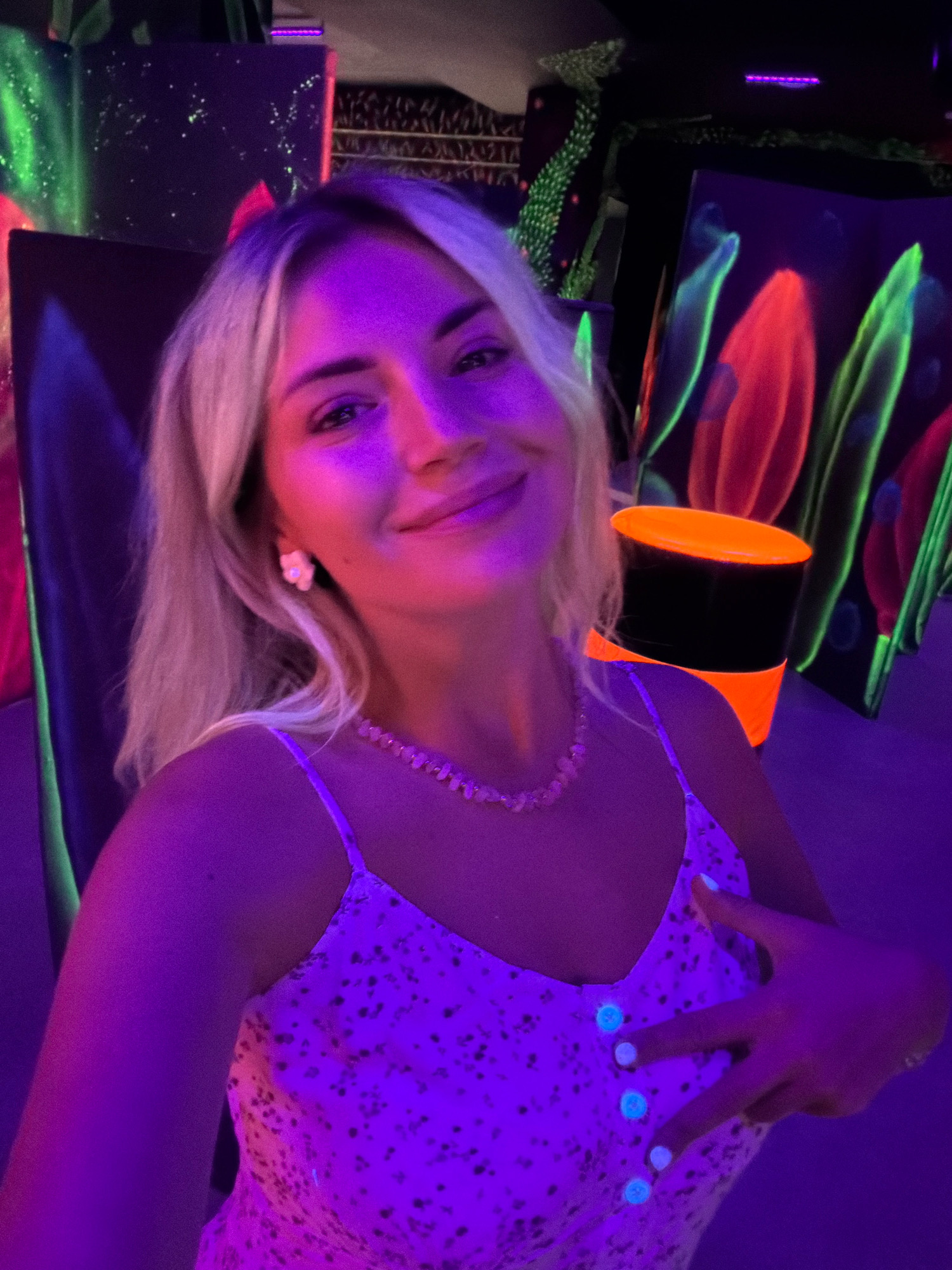 A young blonde woman poses for a selfie in a dimly lit room, illuminated by purple and green neon lights. She is wearing a light-colored dress with a floral pattern and a beaded necklace. Her right hand is raised to her chest, with her fingers forming a peace sign. In the background, there are several large, dark canvases with neon-painted designs of flowers and abstract shapes. To the right, a black cylindrical object with bright orange bands is visible. The overall atmosphere is that of an art installation or an entertainment venue, possibly a glow-in-the-dark art studio or a blacklight mini-golf course. The location of this scene in Podgorica, Montenegro, is not determinable from the visual information provided.
