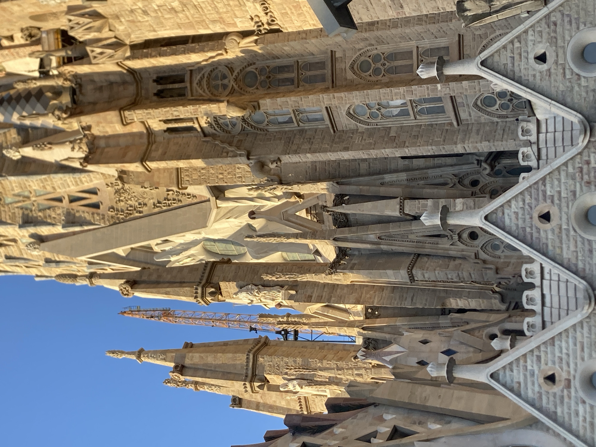 The image shows a close-up, low-angle view of the Sagrada Familia basilica in Barcelona, Spain. The intricate stone architecture is detailed with spires, statues, and ornate windows. A tall orange construction crane is visible rising behind some of the spires on the left side of the image, indicating ongoing construction or renovation. The sky is a clear, bright blue. The sunlight casts warm tones on the stone, highlighting the textures and shadows of the facade. The foreground features the pointed roofs of what appear to be lower structures of the basilica, also adorned with decorative elements.