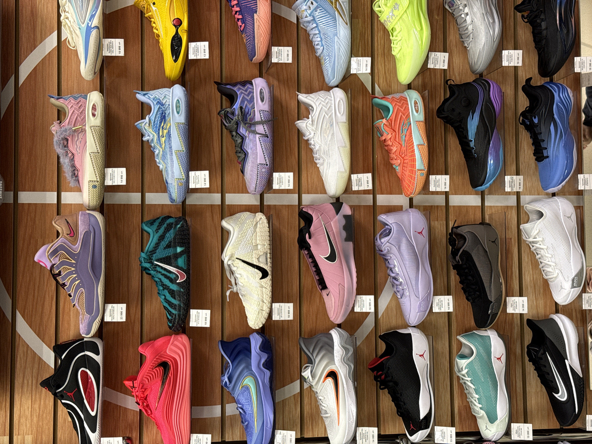 The image shows a wall display of athletic shoes, likely in a store setting. The shoes are various colors and styles, arranged in rows and columns on a wooden background. Price tags are visible beneath each shoe, indicating they are for sale. The location is Krasnoyarsk, Russia.