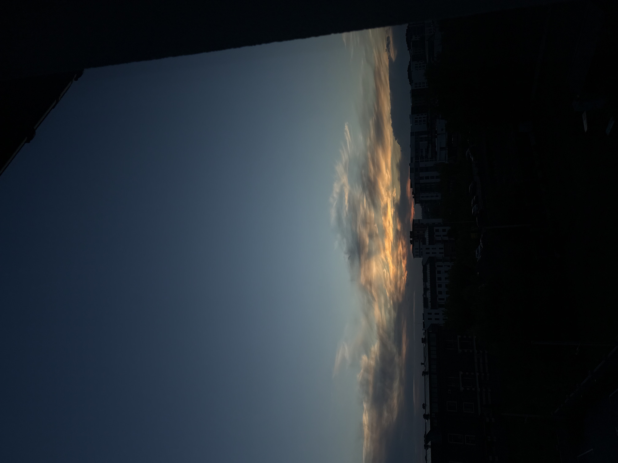 A photograph captures the sky above a city at dusk. The sky is a deep, clear blue, transitioning to hues of orange and yellow near the horizon where a band of clouds is illuminated by the setting sun. These clouds are wispy and spread horizontally, creating a dramatic contrast against the darkening sky. Below the sky, the silhouette of a city skyline is visible. Numerous buildings, including apartments and other multi-story structures, are depicted in dark, indistinct shapes due to the low light. The foreground is almost entirely black, obscuring any details of the ground or immediate surroundings. The image appears to be taken from a vantage point looking out over the city, possibly from a window or balcony, as indicated by the dark, shadowed structure on the right side of the frame.