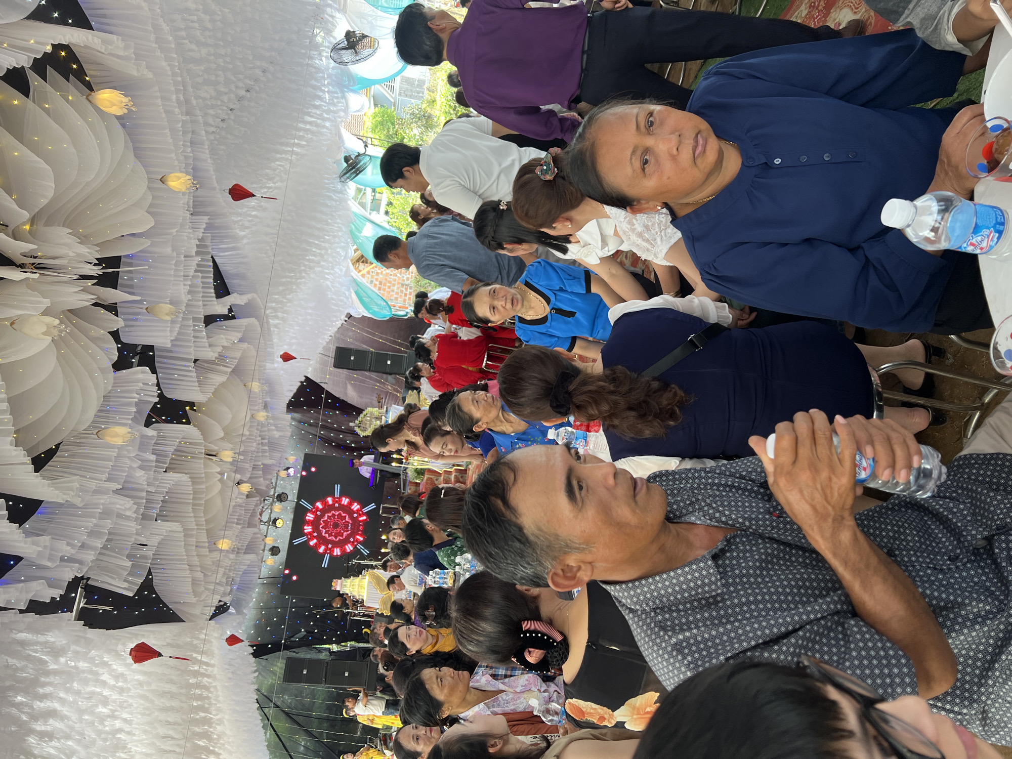 This image captures a vibrant indoor celebration in Bình An, Vietnam, featuring numerous attendees gathered for an event, likely a wedding or a large family reception. The scene is bustling with people of varying ages, predominantly adults, seated at tables arranged for dining.
The venue's ceiling is elaborately decorated with white pleated fabric, resembling layered clouds or petals, interspersed with small warm-toned string lights and larger, flower-shaped decorative lamps. Several red lanterns hang overhead, adding contrasting color. Ceiling fans are visible within the decorated structure.
Towards the back of the room, a stage area is discernible, equipped with large speaker boxes and a prominent circular screen displaying an intricate red and white geometric pattern. A multi-tiered cake is also present near the stage, further suggesting a celebratory occasion.
In the foreground, an older woman in a blue blouse looks directly towards the camera with a neutral expression. To her left, an older man in a patterned short-sleeved shirt holds a "Lavie" branded water bottle, his attention directed towards the stage area. Other guests are scattered throughout the venue, some engaged in conversation, while others observe the activities on stage. Tables are set with plates, cups, and additional "Lavie" water bottles. The overall ambiance appears relaxed and festive, indicating a significant social gathering taking place during what seems to be a well-lit daytime event.