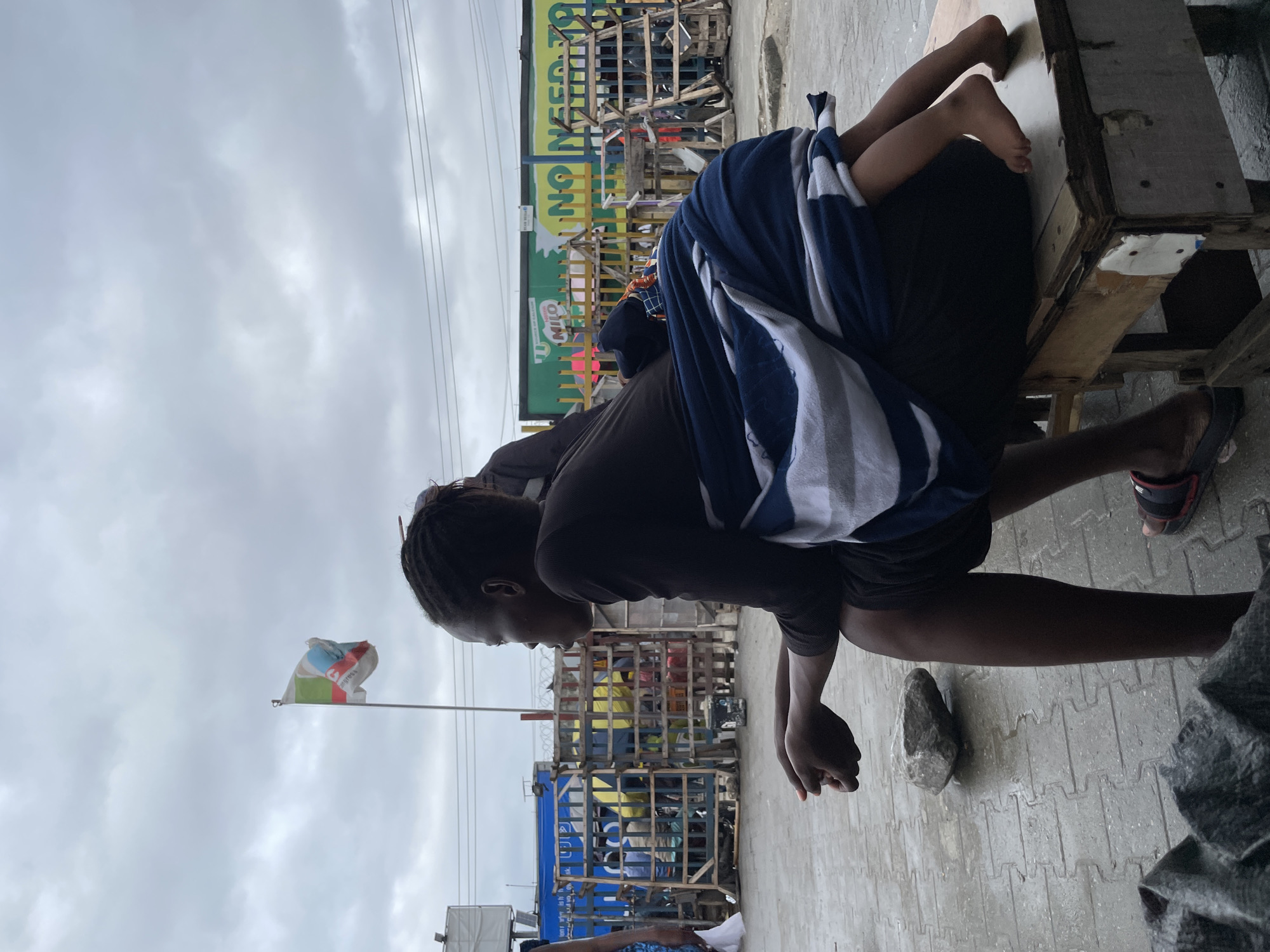 An over-the-shoulder shot captures a woman with braided hair sitting on a wooden stool, facing right, with a child wrapped in a blue and white striped cloth on her back. The woman wears a dark long-sleeved shirt and dark shorts. Her hands are clasped in her lap, resting on a rock. The child's bare legs and feet are visible, dangling over the woman's right side.
The setting appears to be an outdoor marketplace or street in Lekki, Nigeria, indicated by the presence of a Nigerian flag in the background, displaying green, white, and red colors with what looks like a green and blue emblem. Behind the woman, a chain-link fence and wooden barriers separate the foreground from a vibrant green billboard advertising "Milo" and "NO NEED TO TOLL." Power lines are visible above the structures. The ground is paved with interlocking bricks, appearing wet, suggesting recent rain. Other people are partially visible in the background, and various items, possibly market goods, are stacked behind the barriers. The sky is overcast with gray clouds.