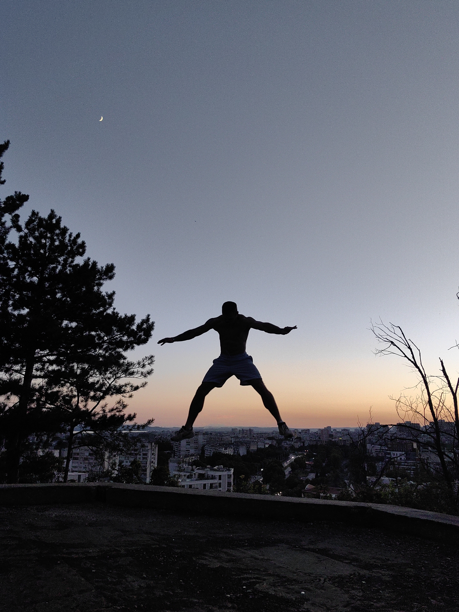 A silhouette of a man is captured mid-air in a dynamic jumping pose against a twilight sky. The man, wearing shorts, is positioned centrally in the frame, with his arms outstretched to his sides and legs spread apart. In the upper left quadrant of the sky, a crescent moon is visible. To the left of the man, a dark silhouette of a pine tree stands prominently. The background reveals a cityscape with buildings and trees, indicative of urban sprawl, with a warm orange and pink gradient painting the horizon where the sun has set. The foreground is dominated by dark, uneven ground, possibly a dirt path or hillside, with a low concrete wall separating it from the city below. The image appears to have been taken from a higher vantage point, looking down upon the city. The location is the Varna Observatory, which is situated on a hill overlooking the city of Varna, Bulgaria.