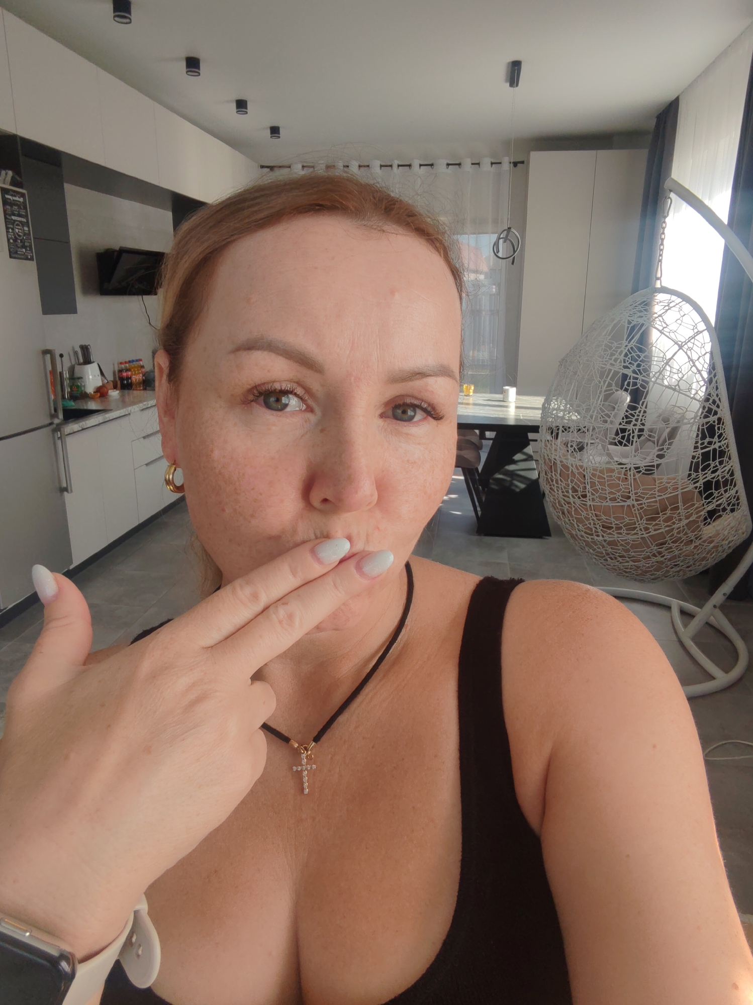 A woman is indoors, likely in an apartment, as evidenced by the presence of a kitchen area with white cabinets and a dining area with a modern table and chairs. She is fair-skinned with reddish-blonde hair, freckles, and blue eyes. She is wearing a black tank top and a necklace with a cross pendant. She is positioned at an angle, appearing to be taking a selfie. Her left hand is raised to her lips in a gesture of kissing or shushing. She is also wearing a smart watch. Behind her is a hanging rattan-style chair. The image was taken in the city of Chaikovsky, Russia.