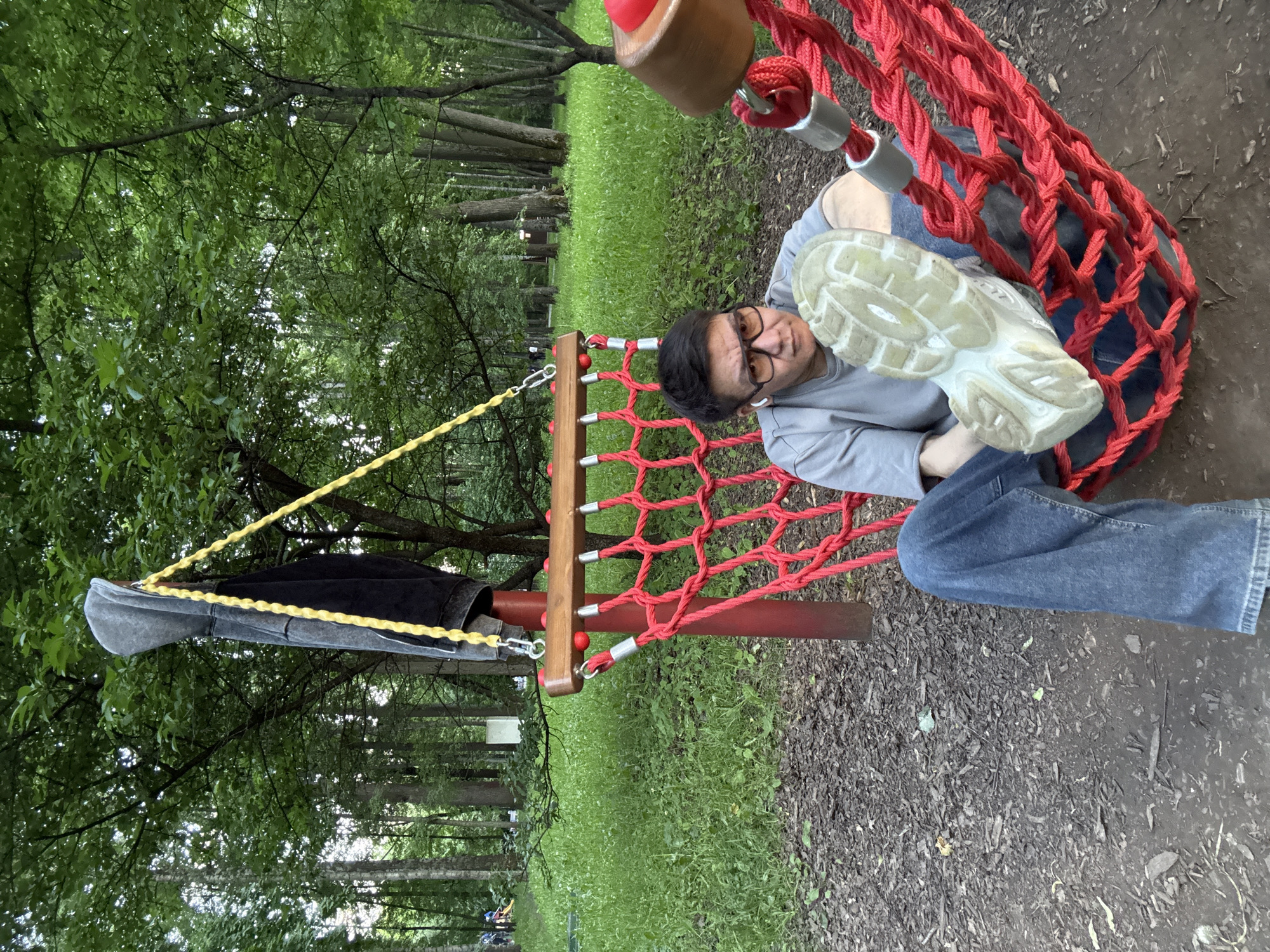A man is depicted relaxing in a red net hammock or swing within an outdoor park setting. He is wearing glasses, a grey long-sleeved shirt, and blue jeans, looking towards the camera with a neutral to slightly amused expression, while holding up the sole of his white sneaker. The hammock features red rope netting, wooden support bars, and metal connectors. A yellow rope or strap extends from one side of the hammock, from which a grey baseball cap and a black jacket are hanging.
The background is dominated by lush green trees and dense foliage, suggesting a wooded or forested area. A vibrant green grassy patch is visible in the middle distance, leading to more trees. The ground in the foreground and beneath the hammock is covered with dark soil or mulch. The lighting is diffused, indicating daytime, possibly under an overcast sky or dense tree cover, contributing to a tranquil atmosphere. No visible text is present in the image. The scene is located in a park in Moscow, Russia.
