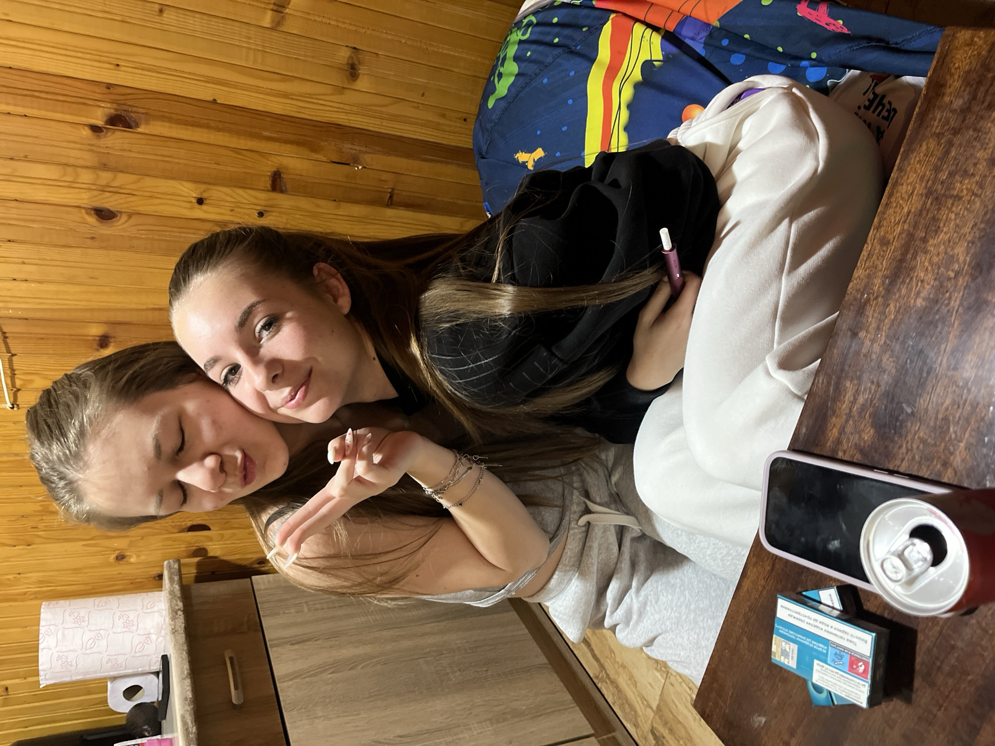 Two young women are close together, appearing to pose for a photo in an indoor setting. One woman has long brown hair, wears a black top, and is seated in white sweatpants. She holds a purple cylindrical object in her hand. The other woman has lighter brown hair and wears a gray tank top. She is touching her fingers together. Both women are fair-skinned. A phone, an open can of a dark beverage, and a blue box are on a wooden table next to them. A colorful blanket sits in the background. The walls are wooden. There is no specific indication of Nova Zagora, Bulgaria, in the image.
