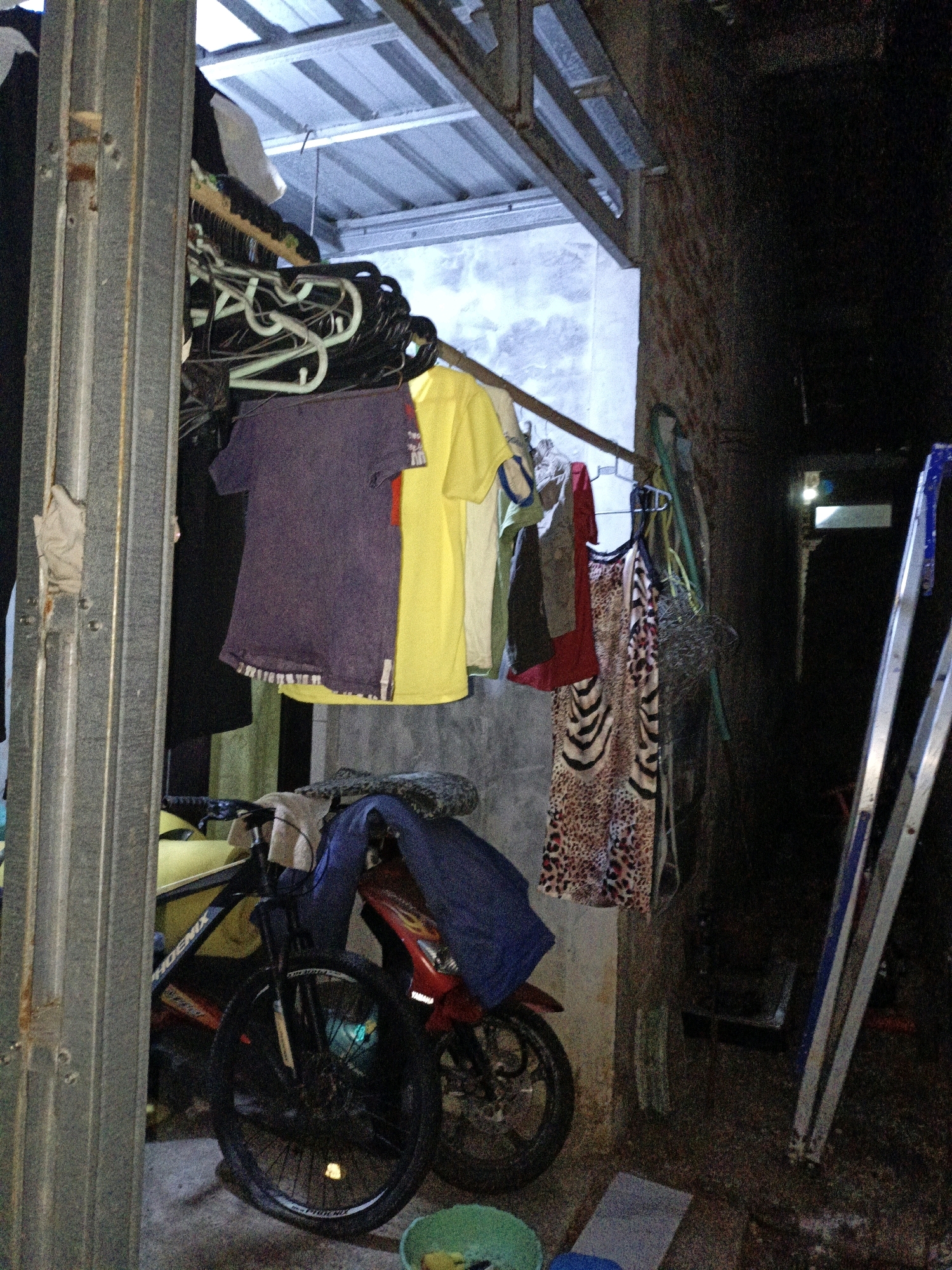 In an outdoor or semi-enclosed area in Cikal, Indonesia, a bicycle with a black frame and black tires is parked. The bicycle has a blue cover draped over its handlebars and seat. To the right of the bicycle, a motorcycle with a red body is partially visible. Clothing is hanging on a clothesline that extends horizontally across the scene. Among the hanging clothes are a purple t-shirt, a yellow t-shirt, and a patterned dress. A silver ladder leans against a brick wall on the right side of the image. The ground appears to be concrete or dirt. The overall impression is a cluttered utilitarian space.