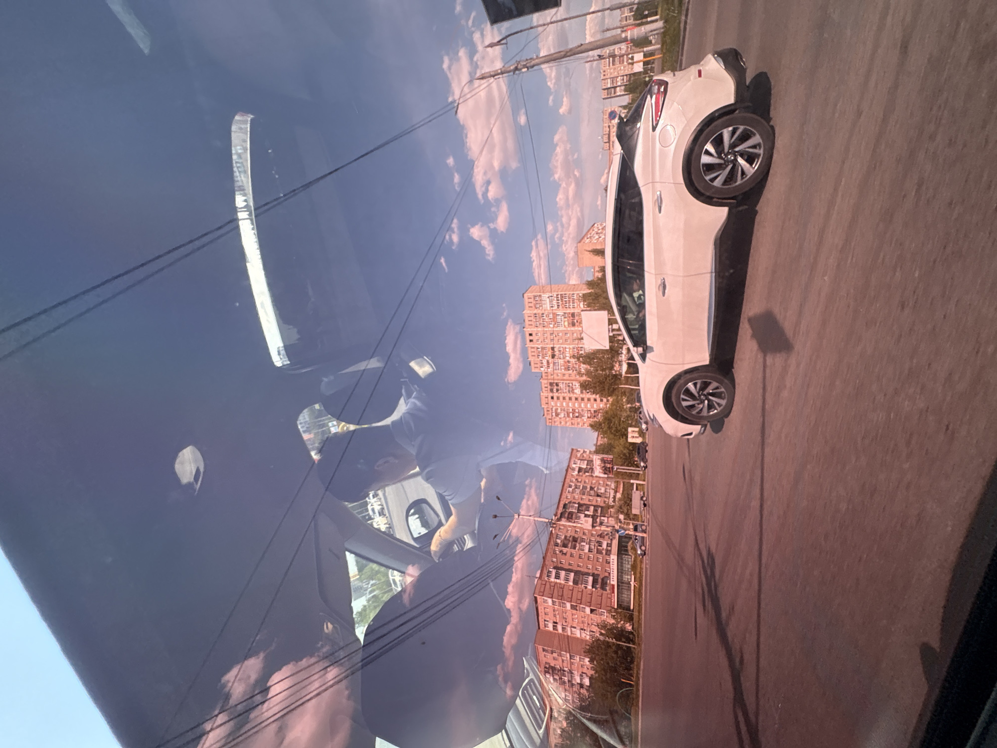 The image is taken from inside a car, looking out of the windshield. The driver is visible in the reflection on the glass. The camera is pointed toward another car, which is white, on a paved road running through an urban environment. The street is lined with multiple multistory apartment buildings, a common architectural feature of Izhevsk. The sky is blue with white clouds. Electrical lines are visible in the space between the camera car and the white car, above the street. Based on the architectural style of the buildings visible in the background, the location is likely Izhevsk, Russia.